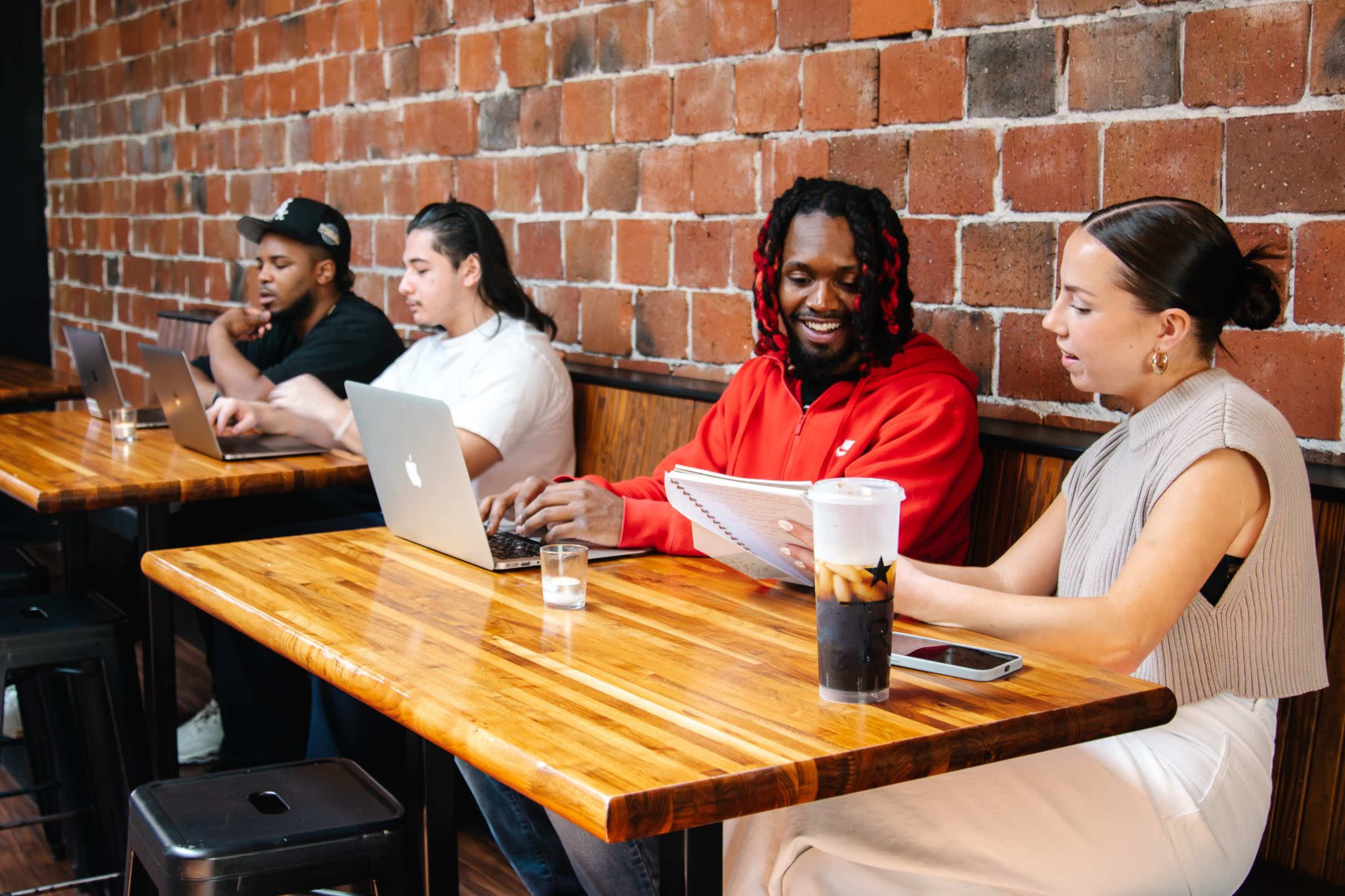 Four individuals work on laptops at a wooden table in a café, with one person reviewing notes and another sitting with a drink.