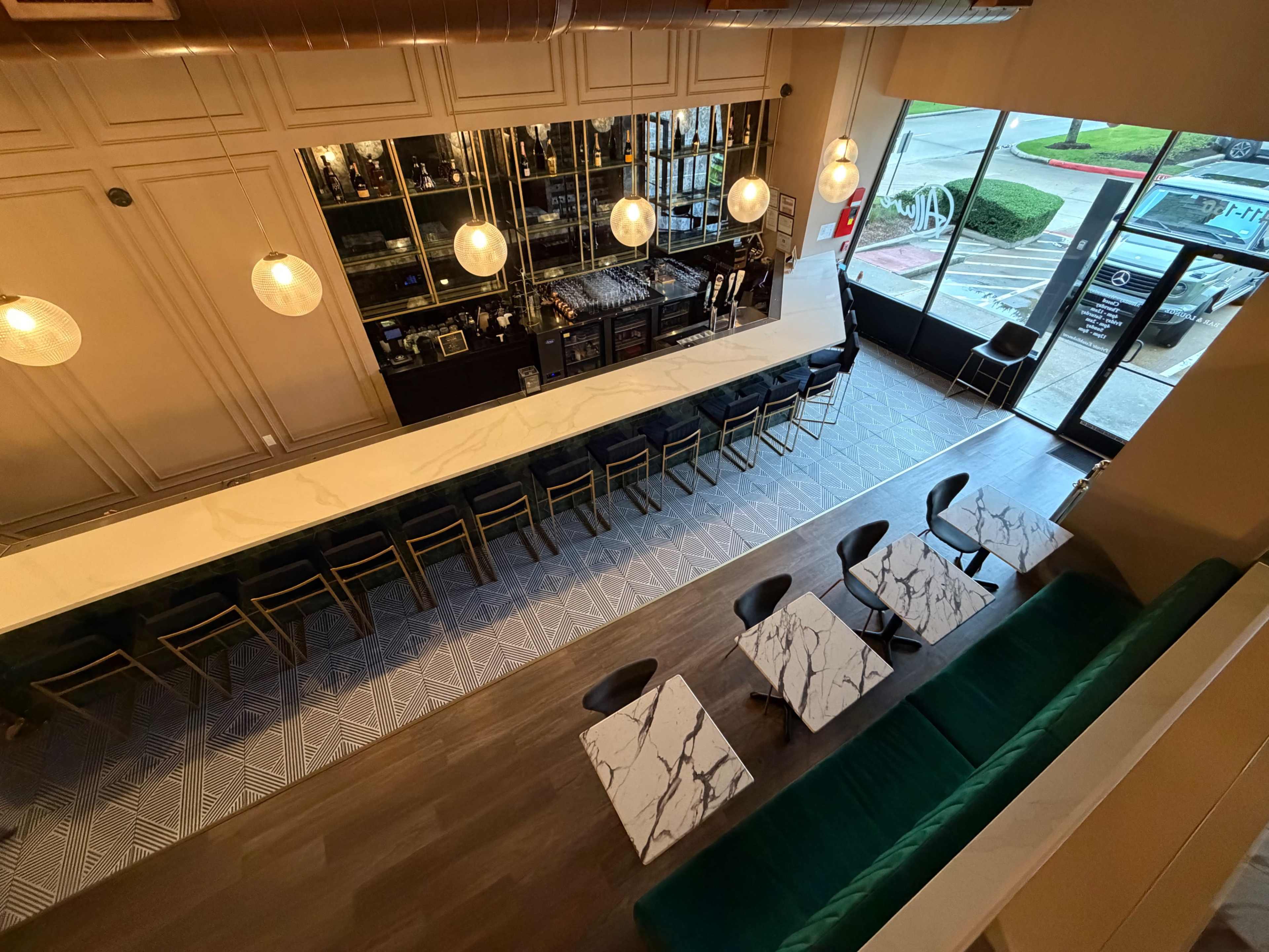 The image shows an overhead view of a modern bar area with a long marble counter, green seating, and several tables with marble tops.