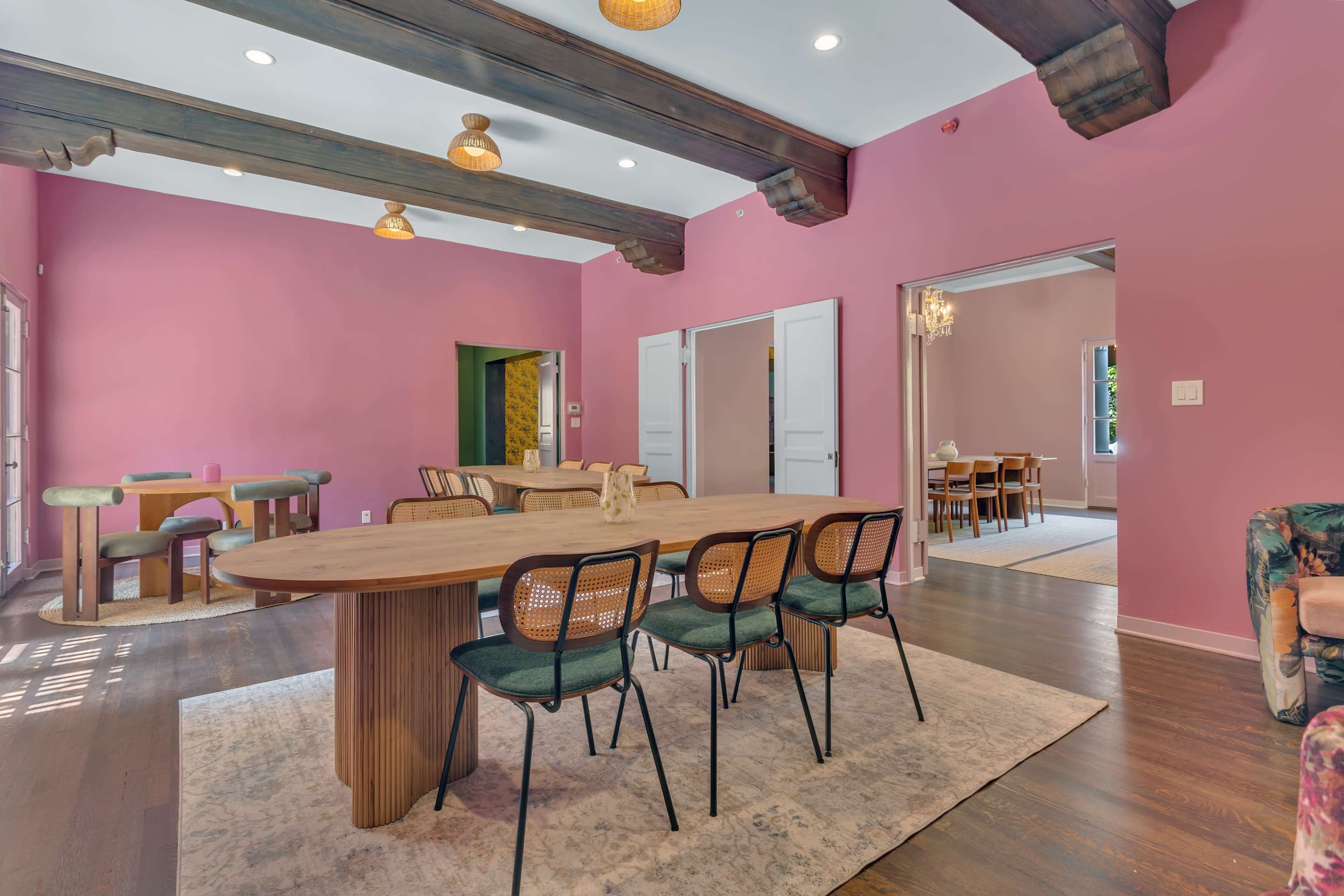 A spacious dining area features a long wooden table surrounded by black chairs, with pink walls and exposed wooden beams overhead.
