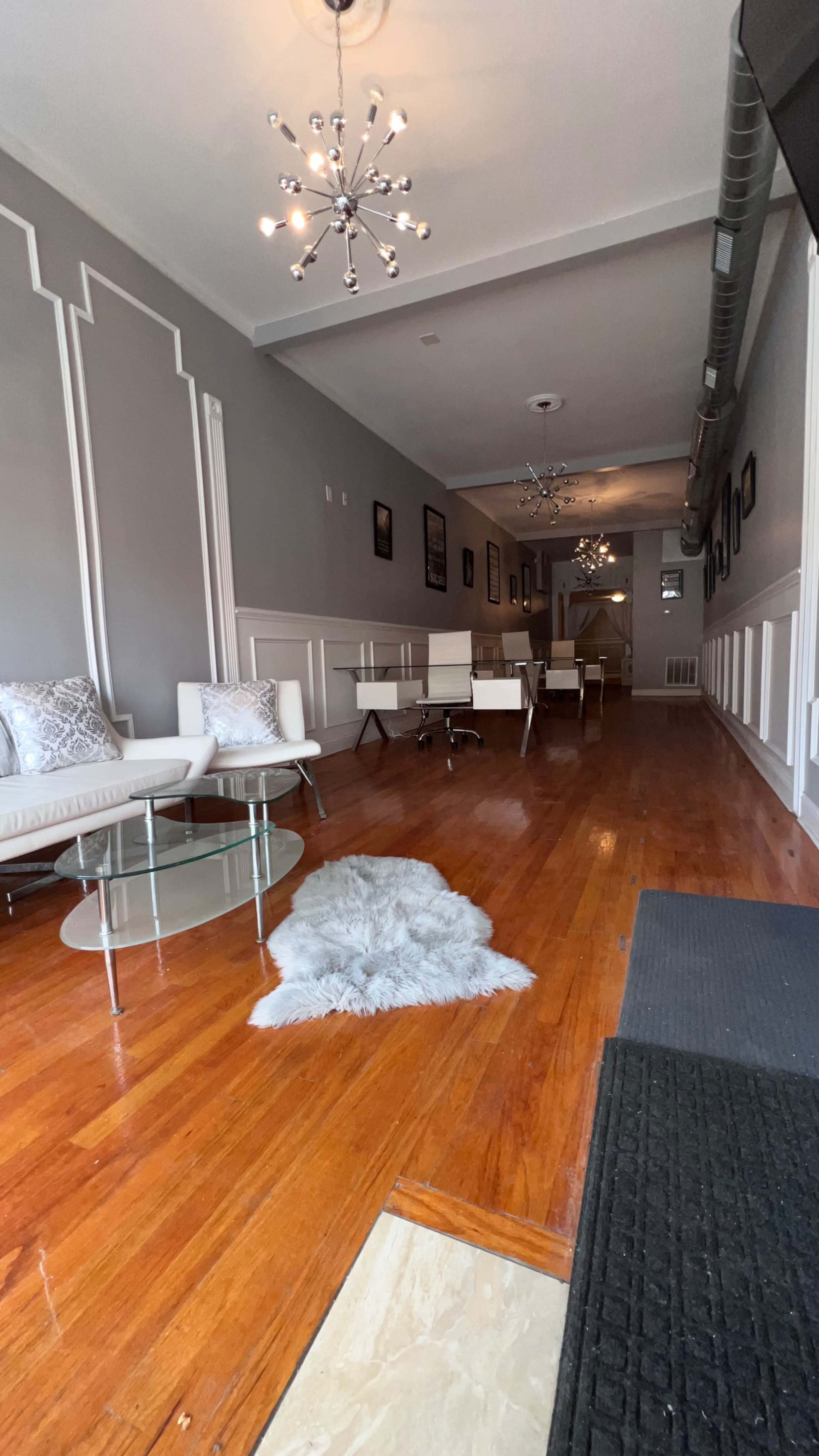 The image shows a modern interior of a room with wooden floors, white furniture, a glass table, and multiple light fixtures, featuring a long hallway leading to another area.