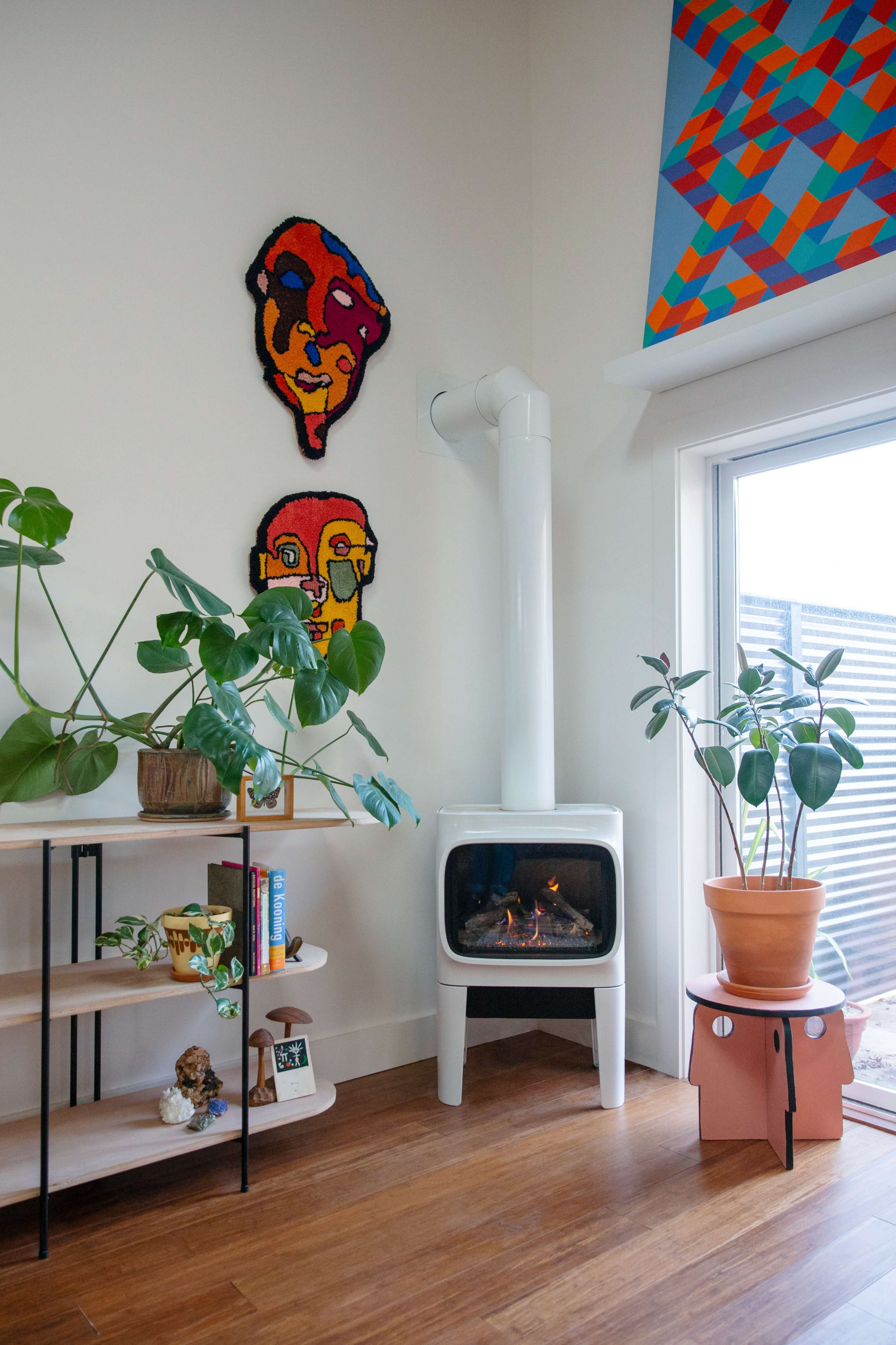 A cozy living room corner featuring a white wood stove, plants on a shelf, and colorful wall art above.