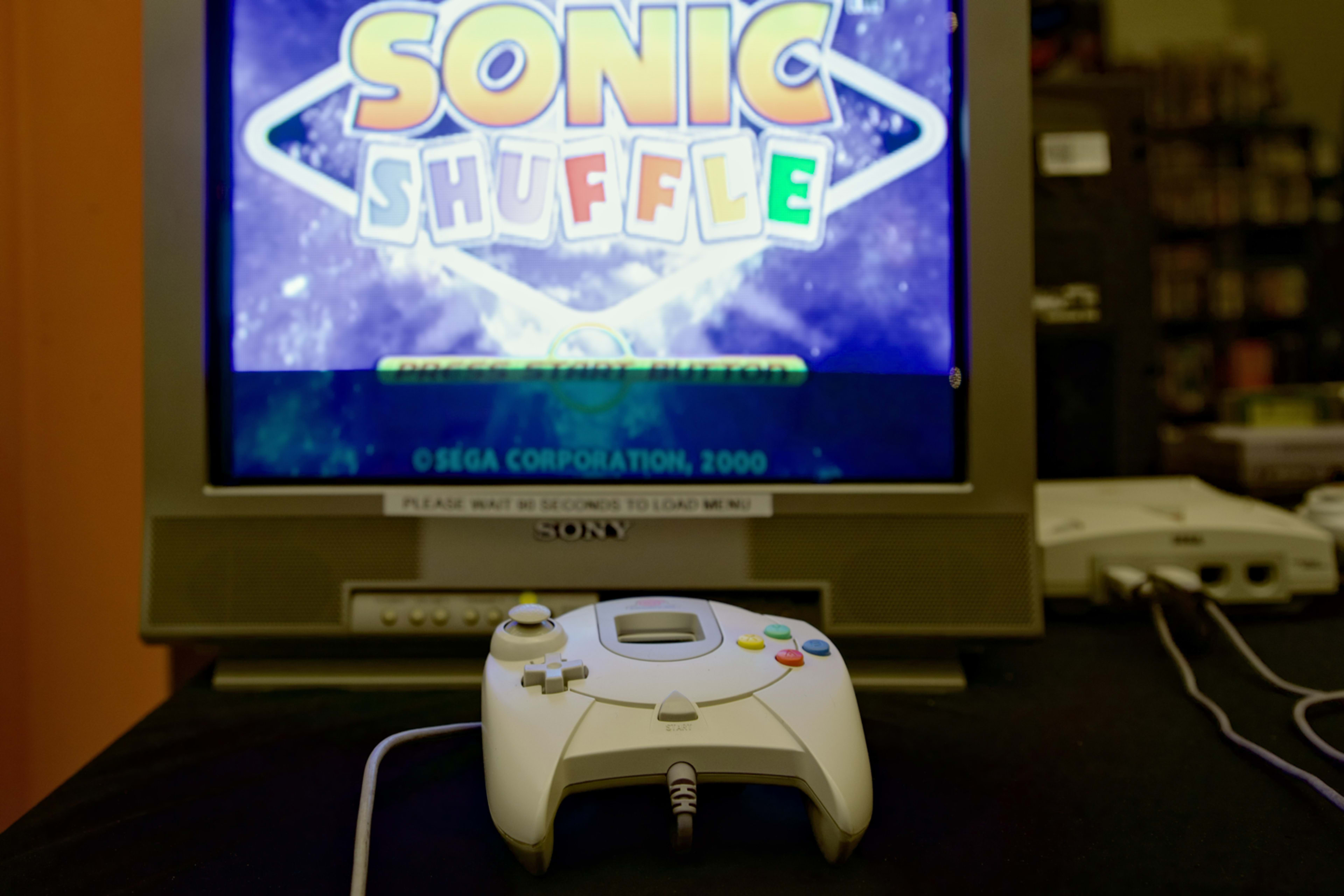 A Sega Dreamcast controller sits in front of a television displaying the title screen for "Sonic Shuffle."