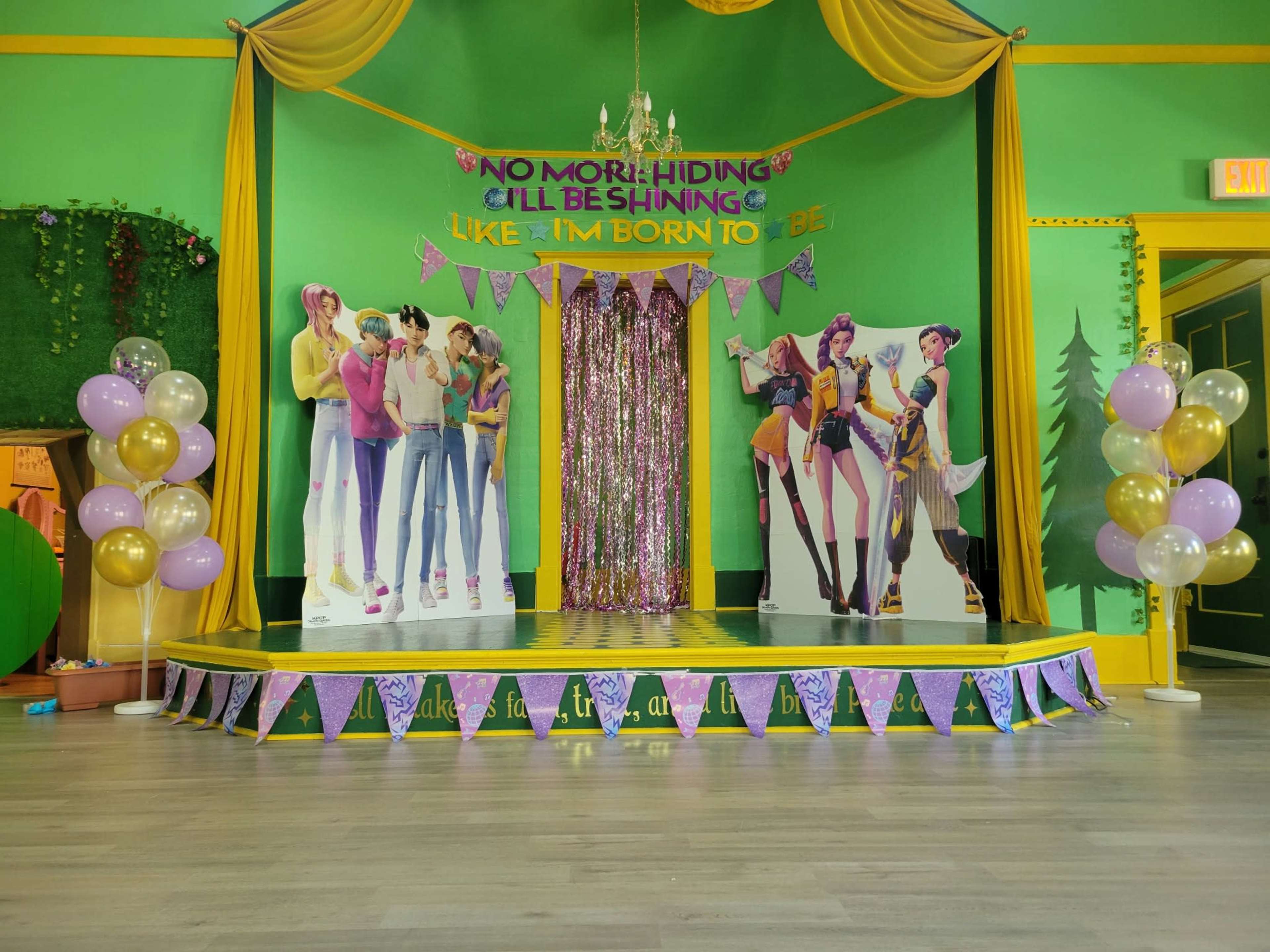 The image shows a brightly decorated stage with green walls, yellow drapery, and vibrant banners, featuring lifelike cutouts of characters and colorful balloons.