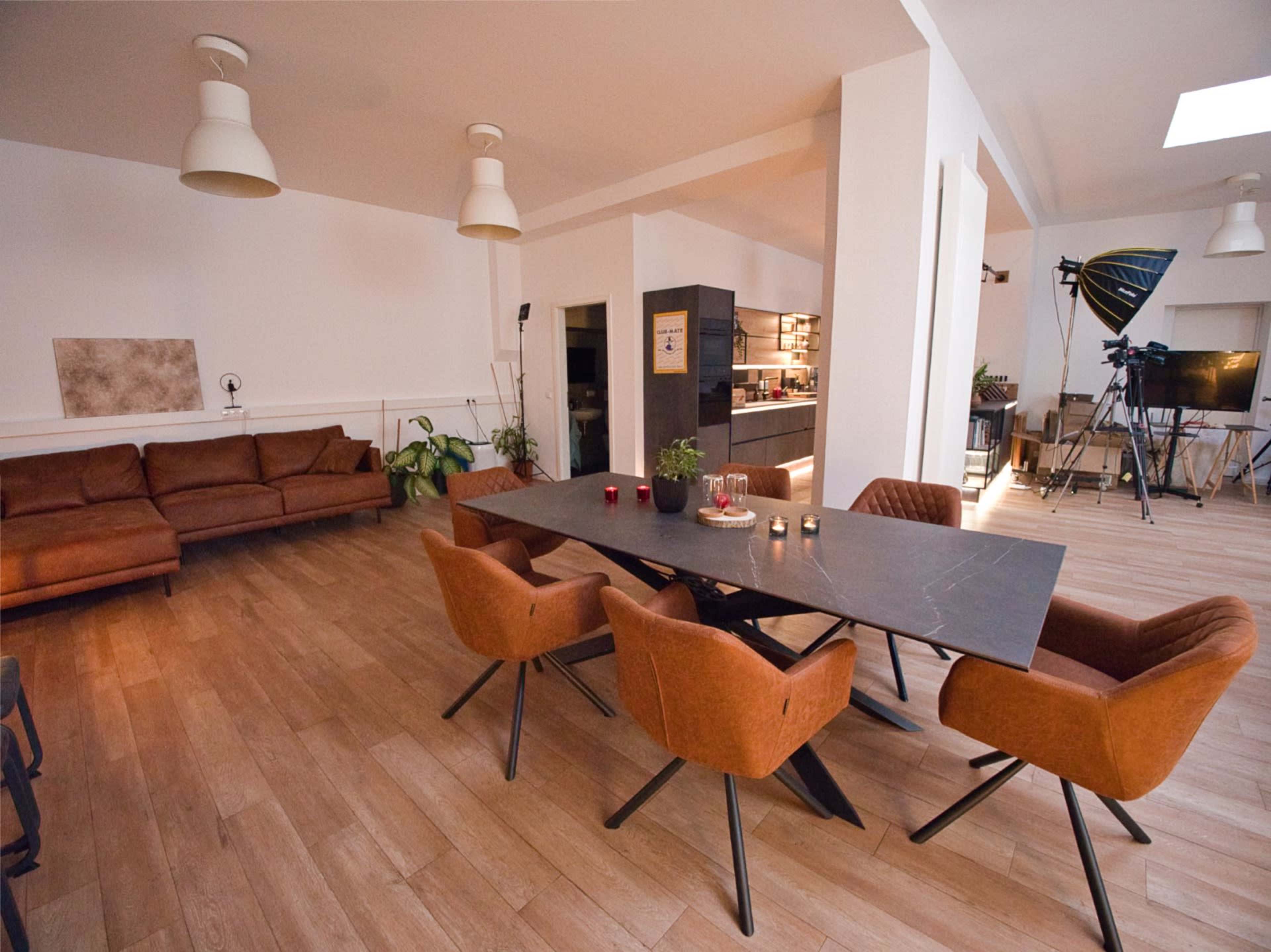 The image shows a spacious, modern living area with a large dining table surrounded by orange chairs, a brown sofa, and a well-lit kitchen in the background.