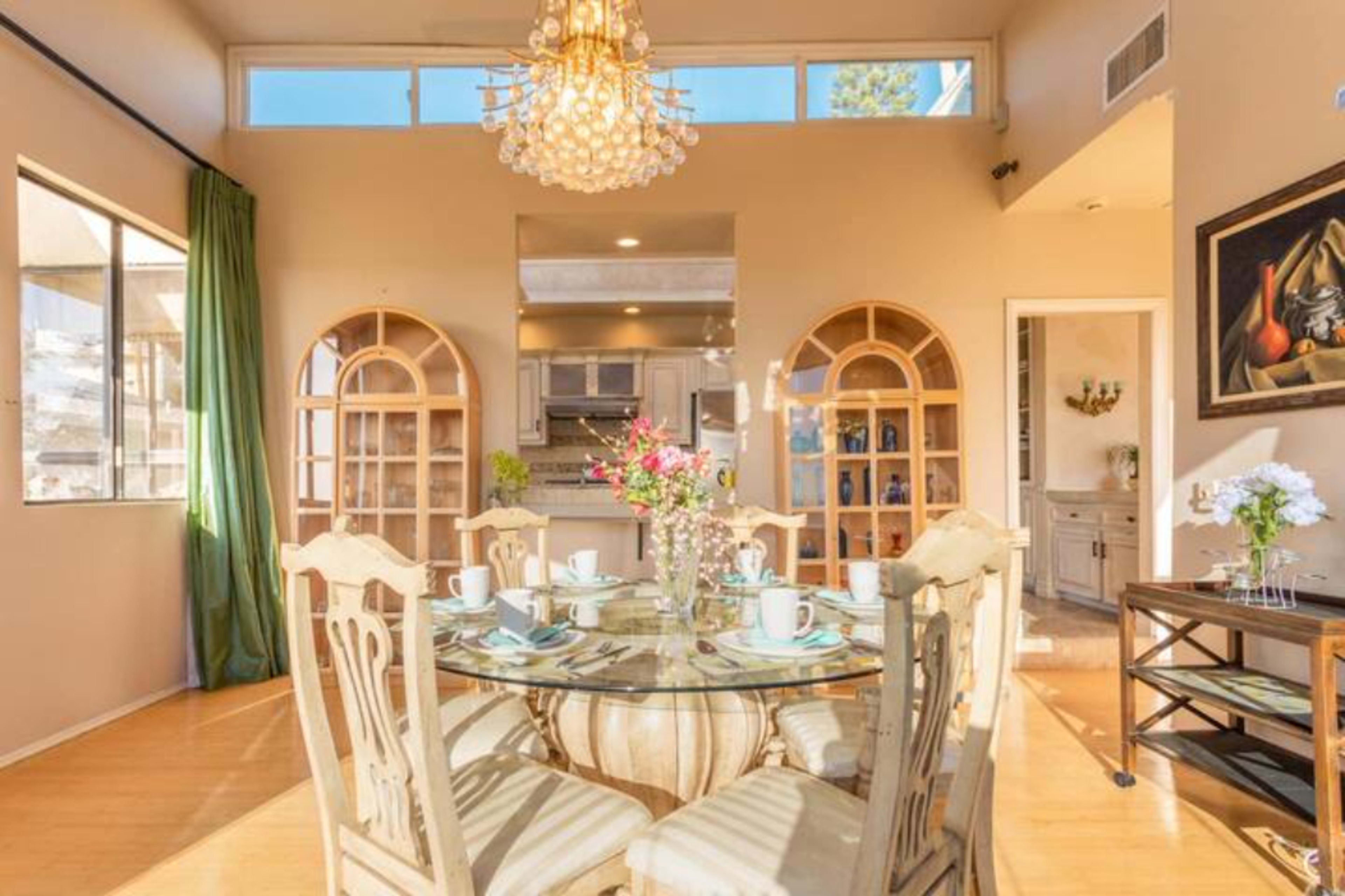 A dining area featuring a round glass table surrounded by six wooden chairs, with a chandelier overhead and kitchen visible in the background.