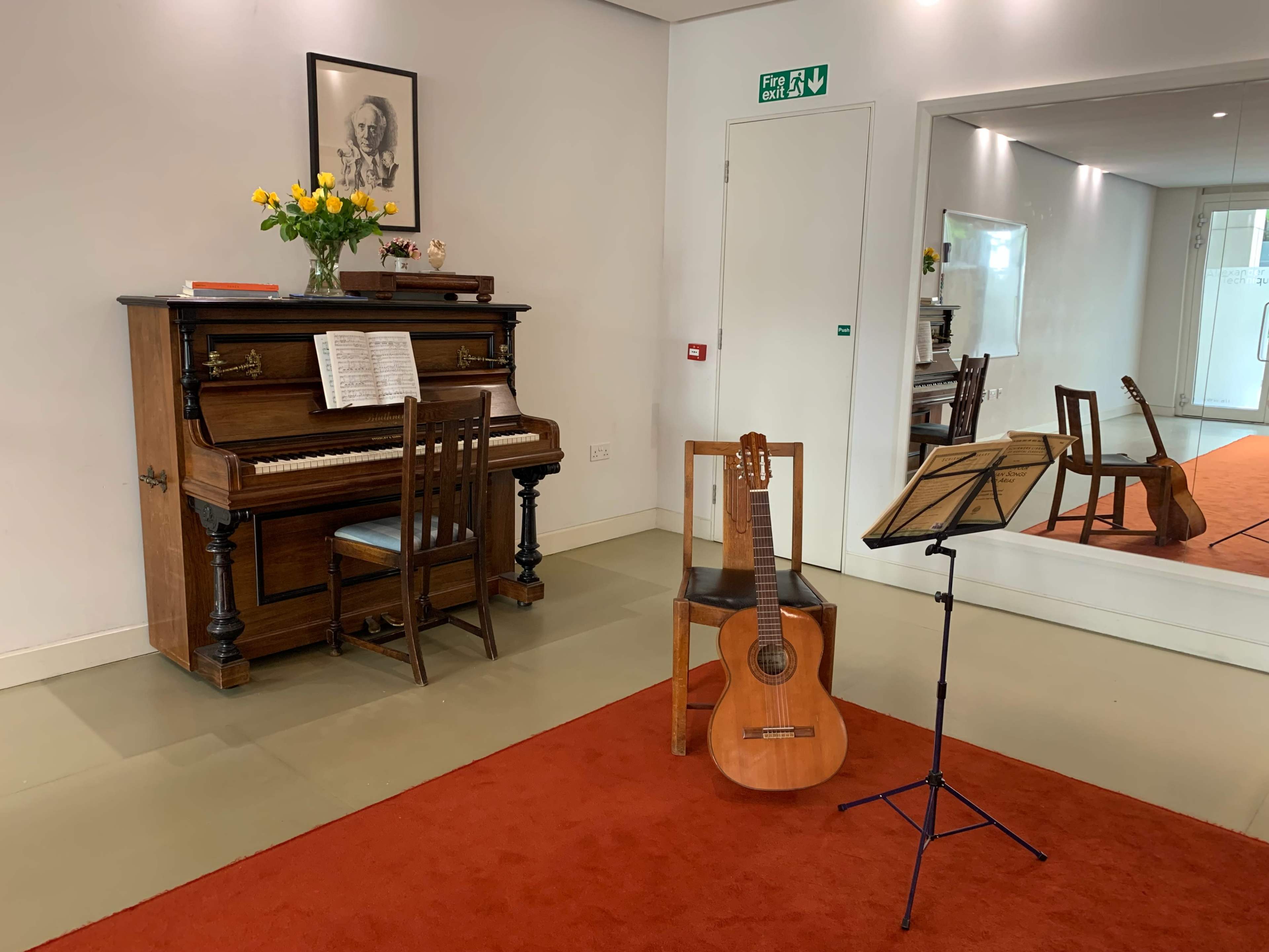The scene features an upright piano beside a wooden chair, with a guitar and a music stand placed nearby on a red carpet in a well-lit room.