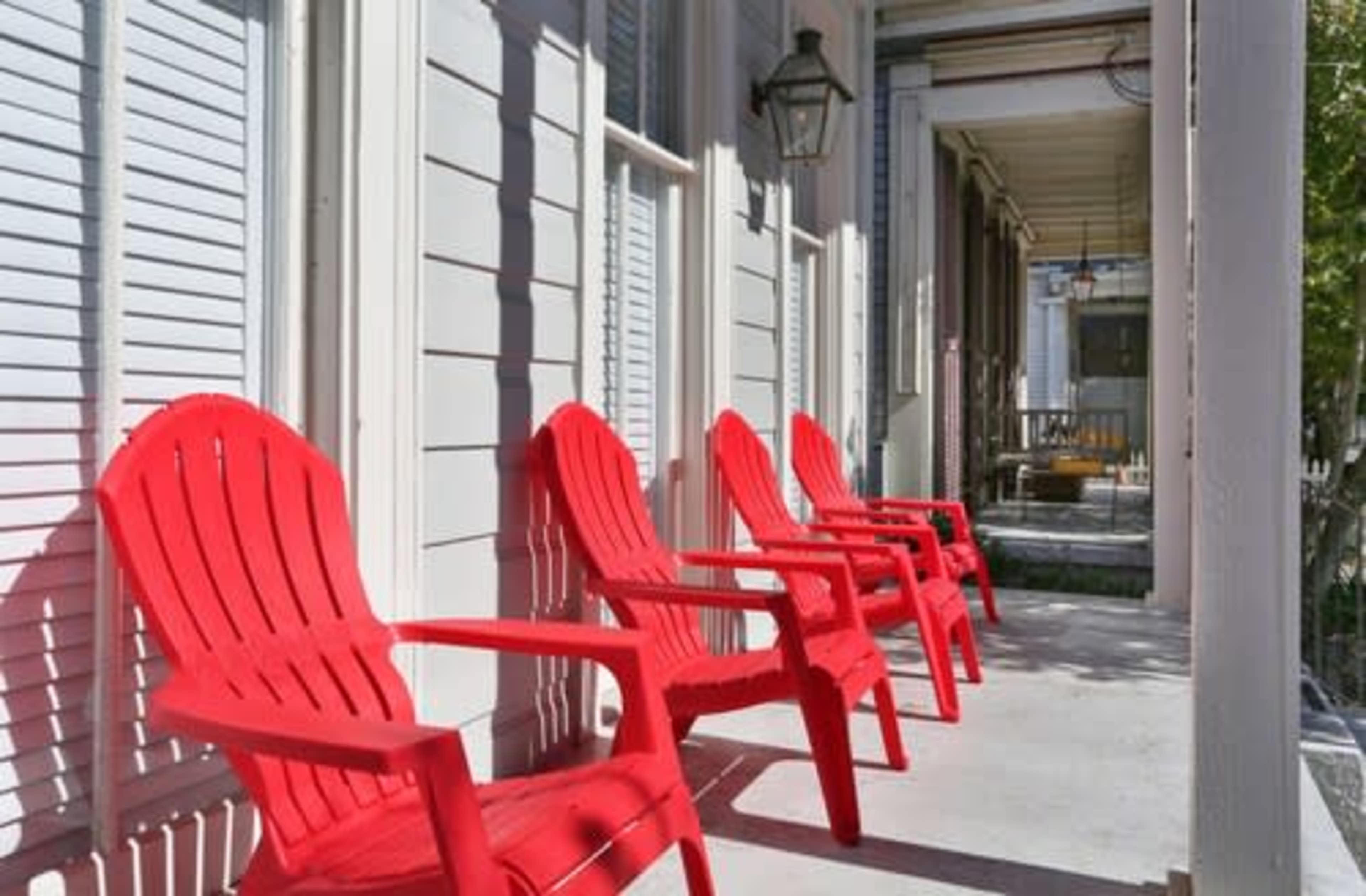 Four red Adirondack chairs are arranged along a white porch with shuttered windows.
