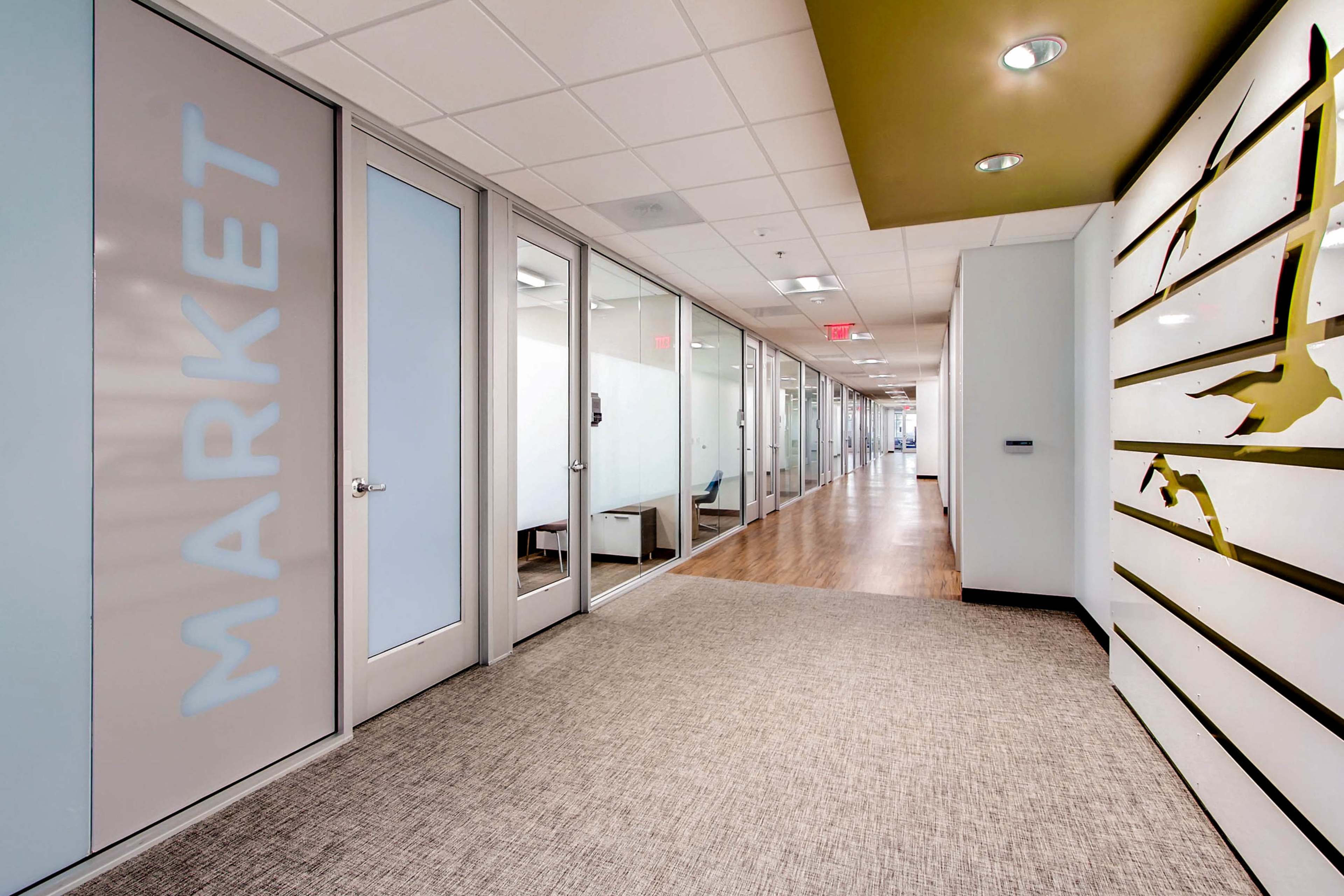 A modern office hallway lined with glass-walled conference rooms on one side and a textured wall with decorative elements on the other.