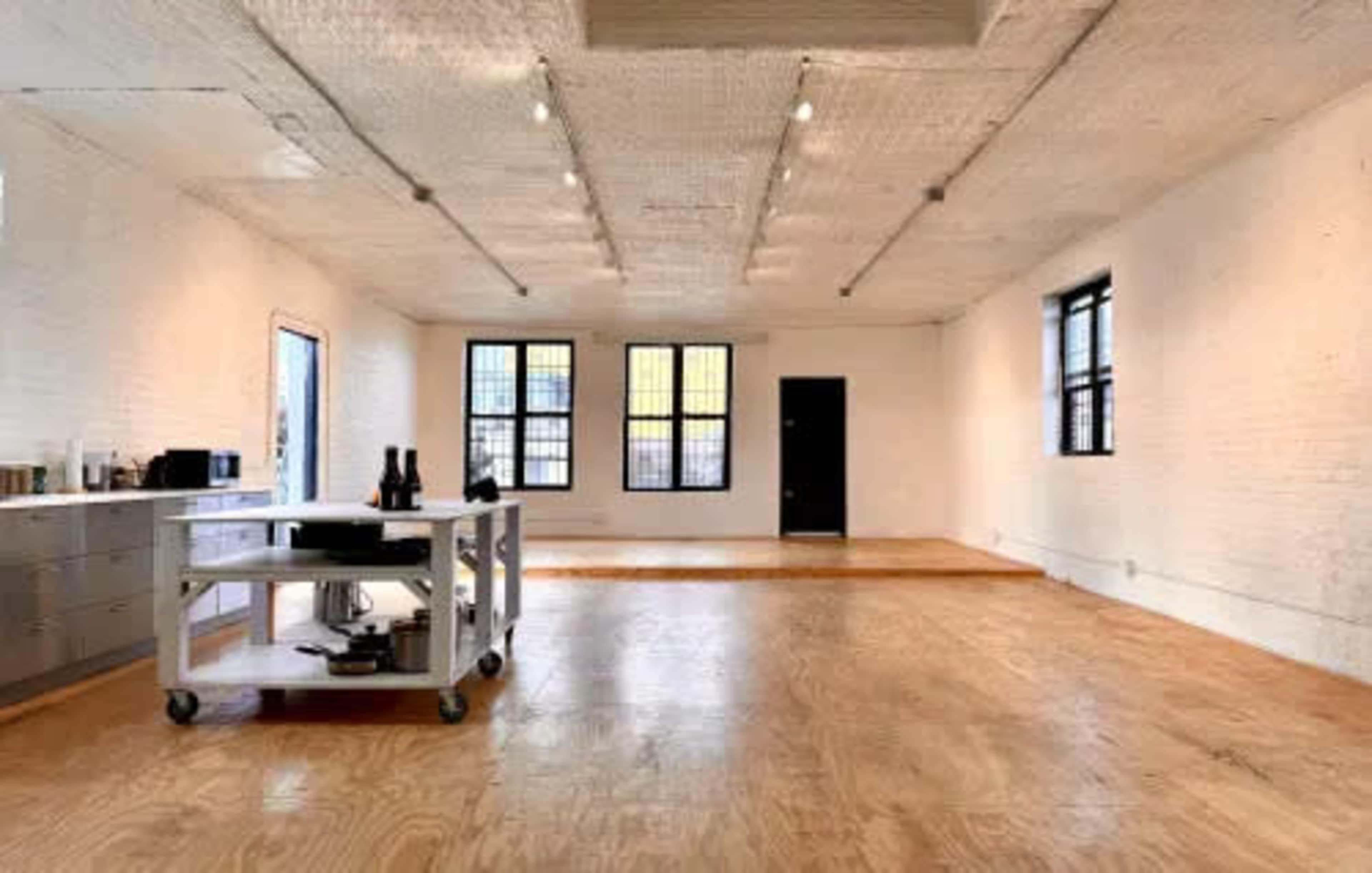 The image shows a spacious, empty room with wooden flooring, white walls, and a rolling cart with kitchen items on one side.