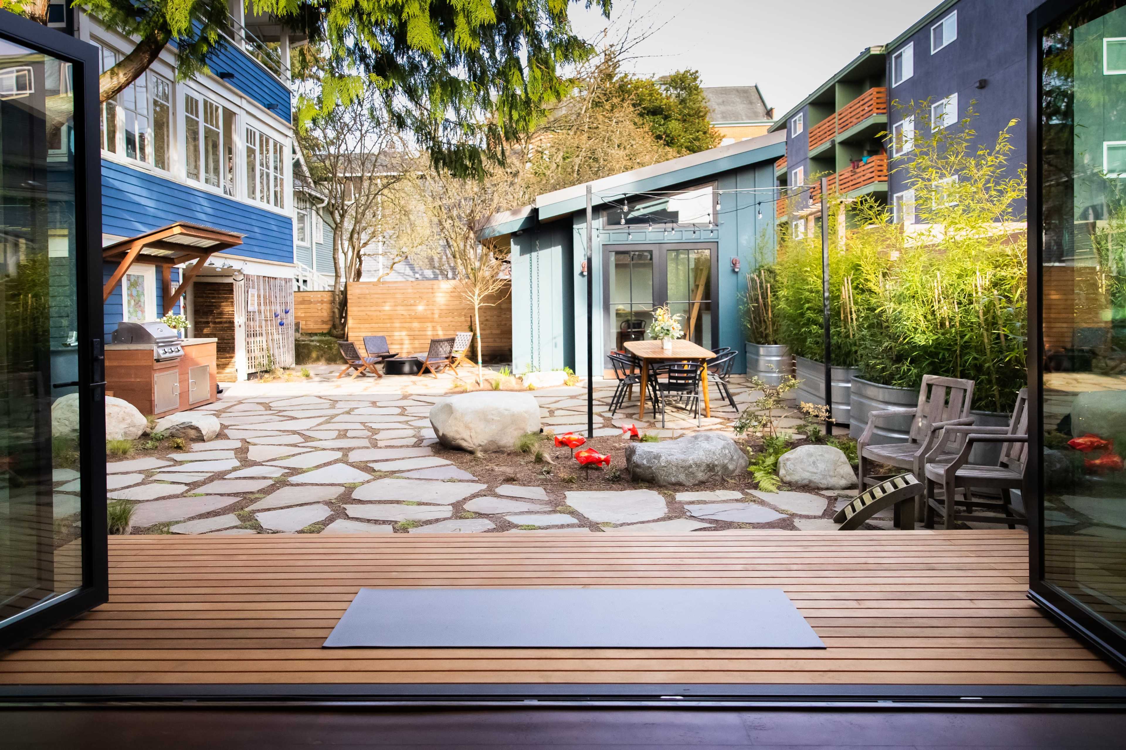 The image shows a modern outdoor courtyard with stone pathways, a wooden deck, and a mix of seating areas surrounded by greenery and buildings.