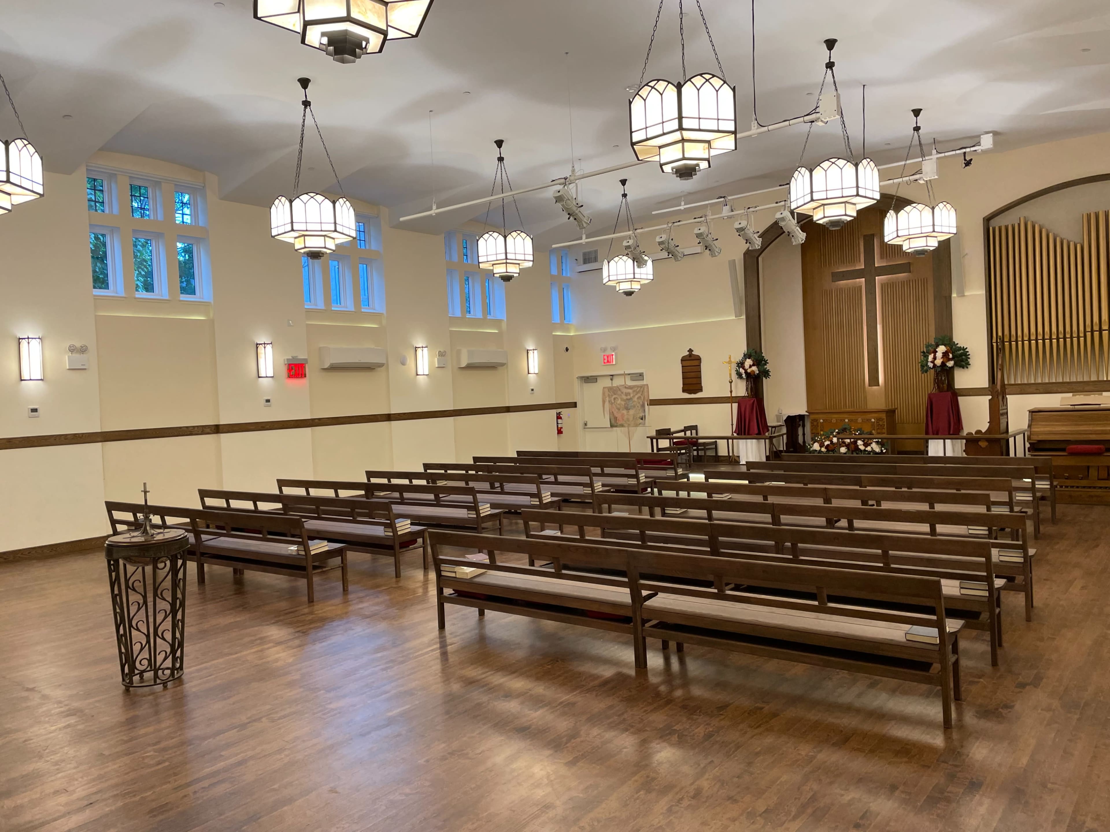 An empty church interior with wooden pews arranged in rows, a wooden stage at the front, and decorative lighting fixtures hanging from the ceiling.