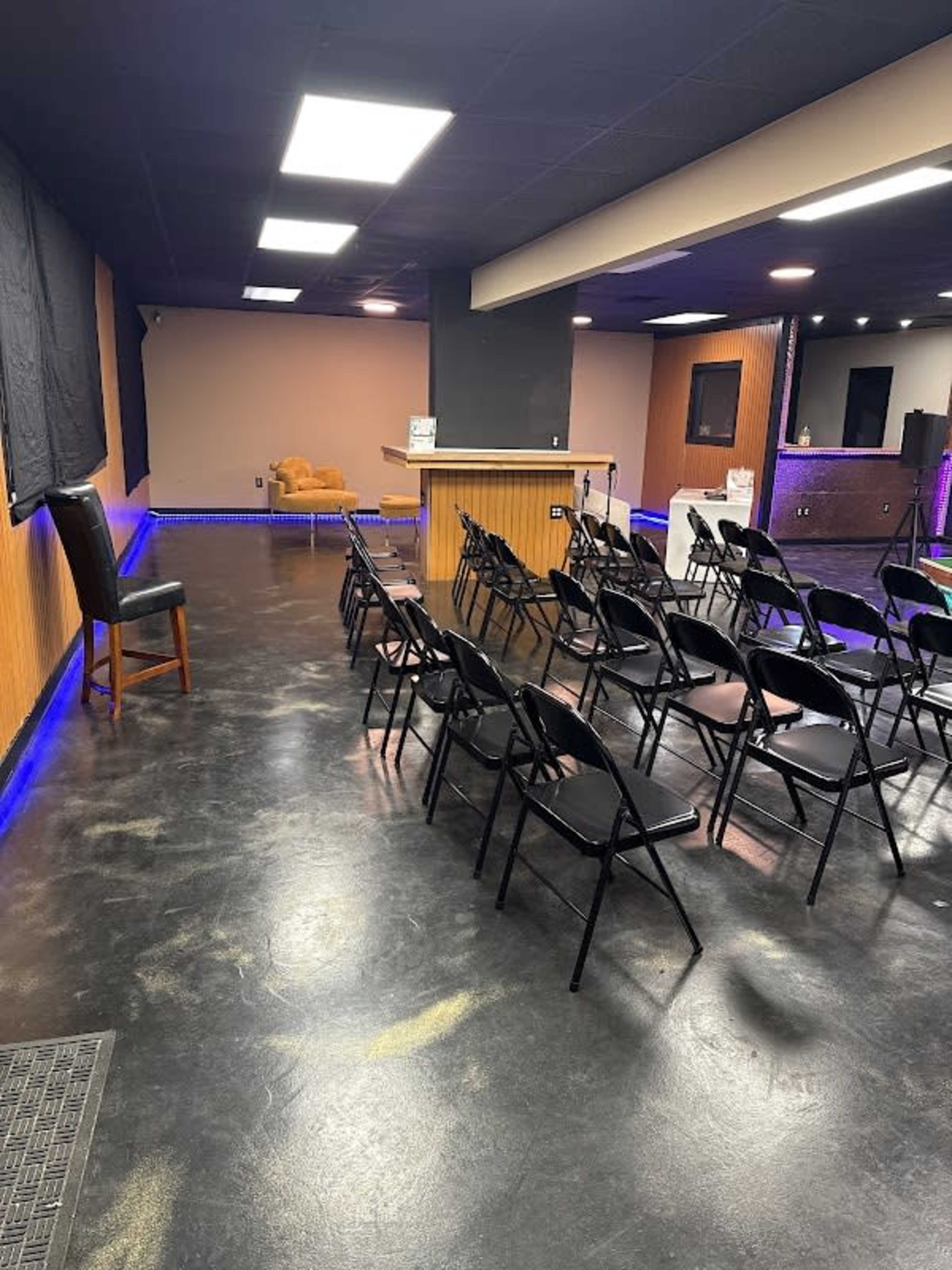 The image shows a room set up with rows of black folding chairs facing a small bar area, with a plush seating arrangement in the background and purple lighting along the floor.