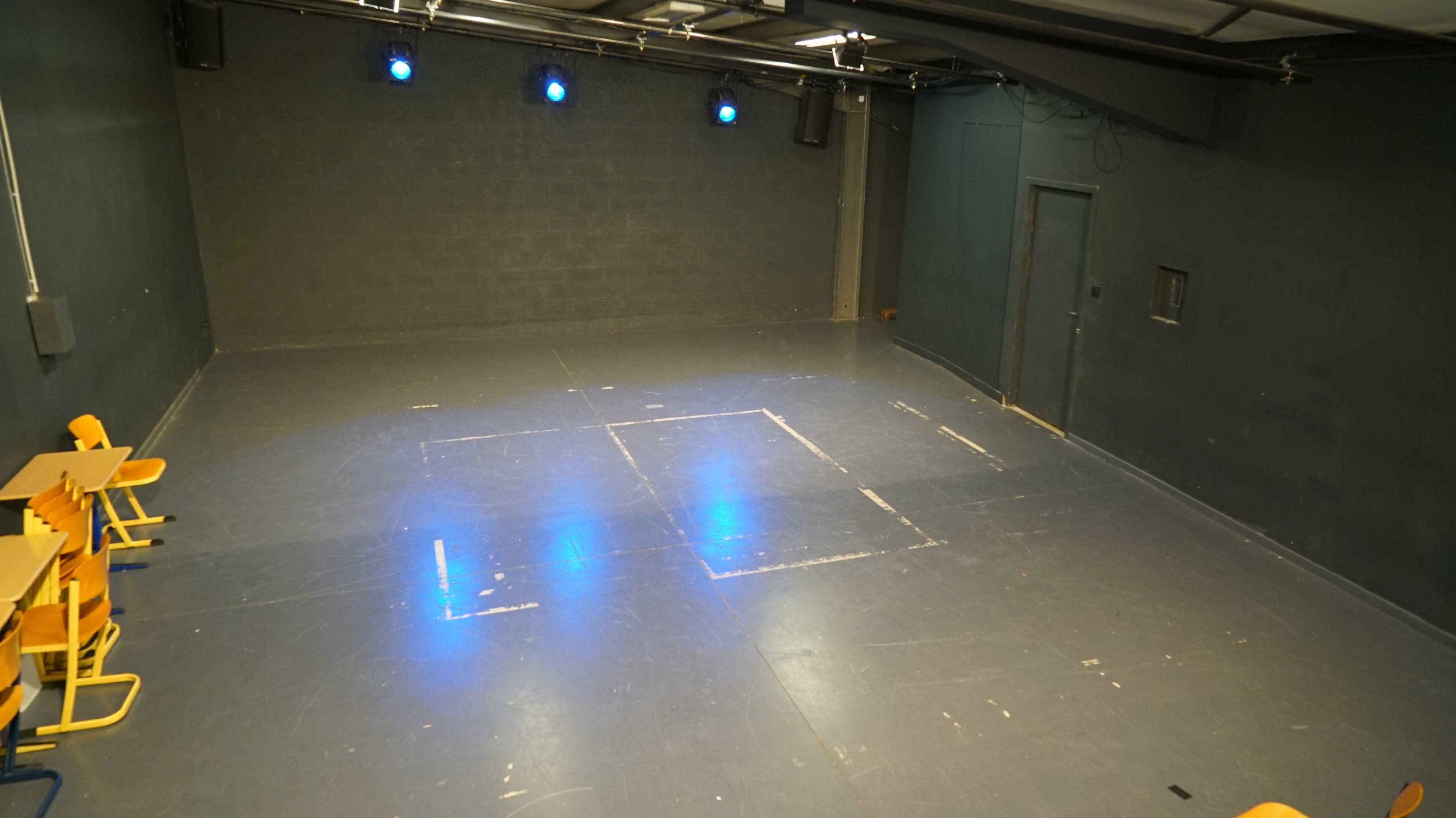 A sparsely furnished rehearsal space with a bare floor and blue stage lights illuminating the area.