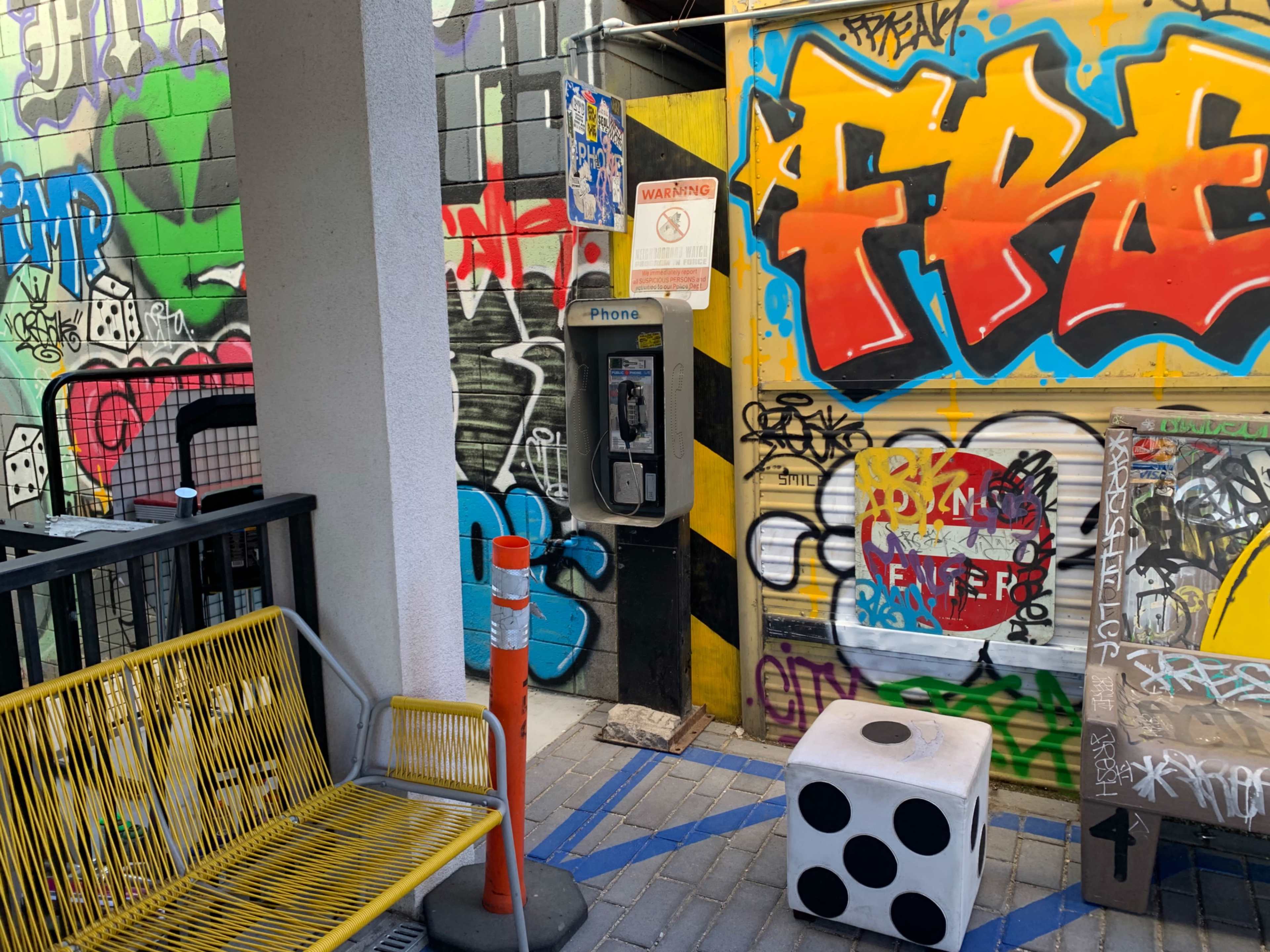 A payphone stands against a graffiti-covered wall in an alley, with a yellow bench and a large white die nearby.