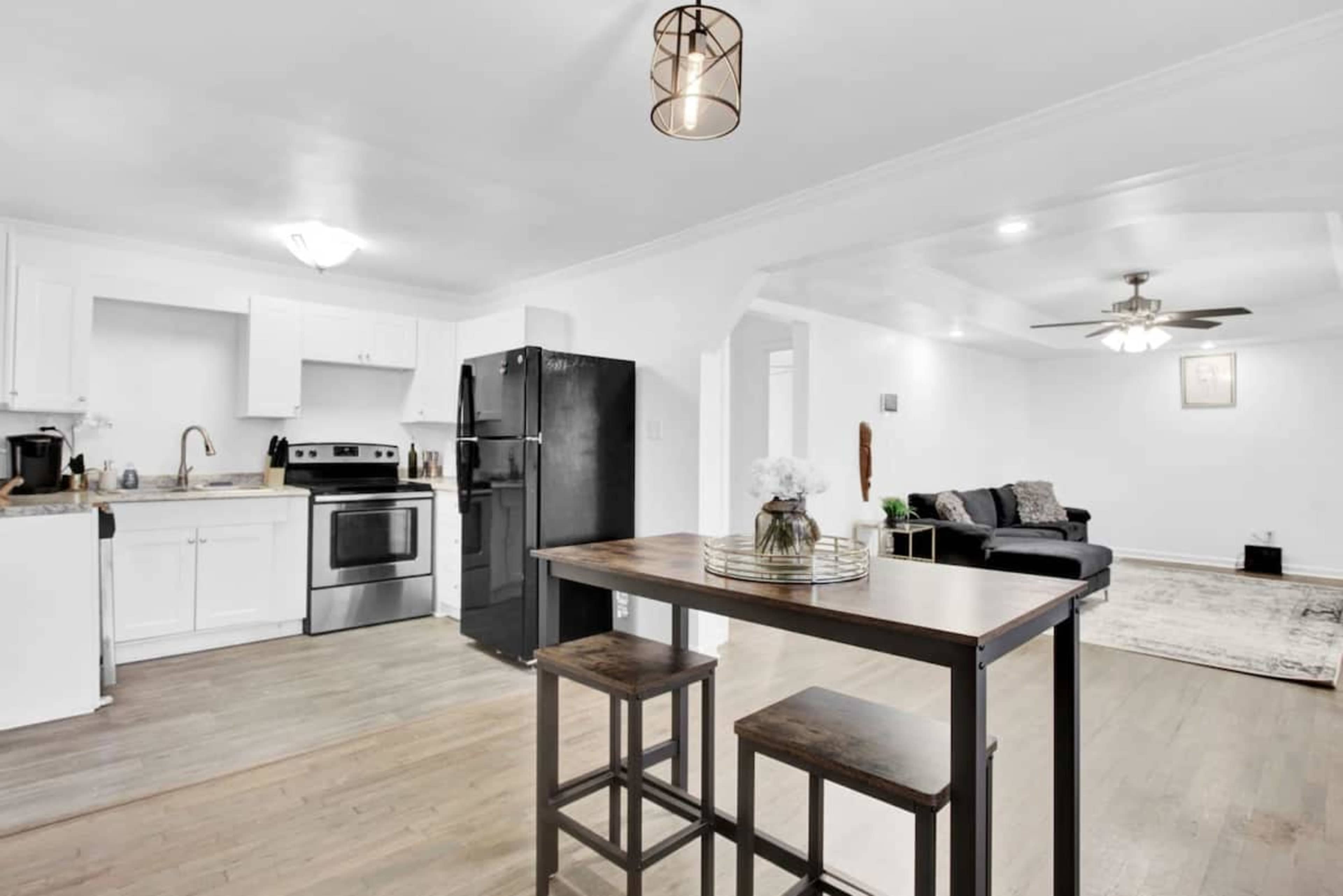 A modern kitchen and living area with white cabinetry, stainless steel appliances, and a wooden dining table with stools.