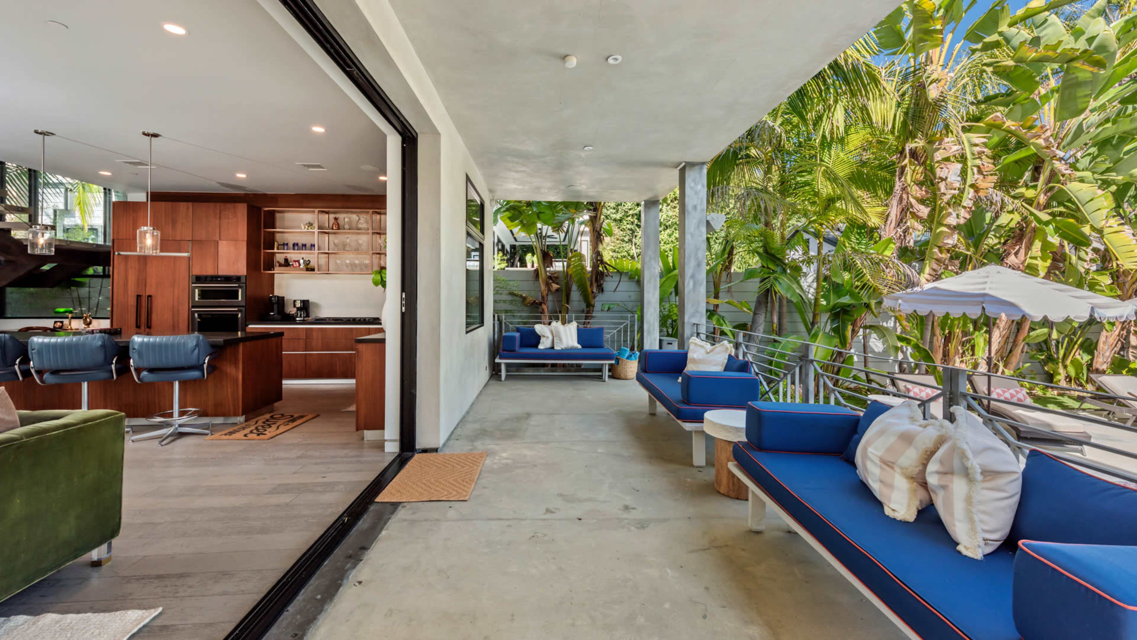 The image shows a modern outdoor patio area furnished with blue seating and a small table, adjacent to a contemporary kitchen that features wooden cabinetry.