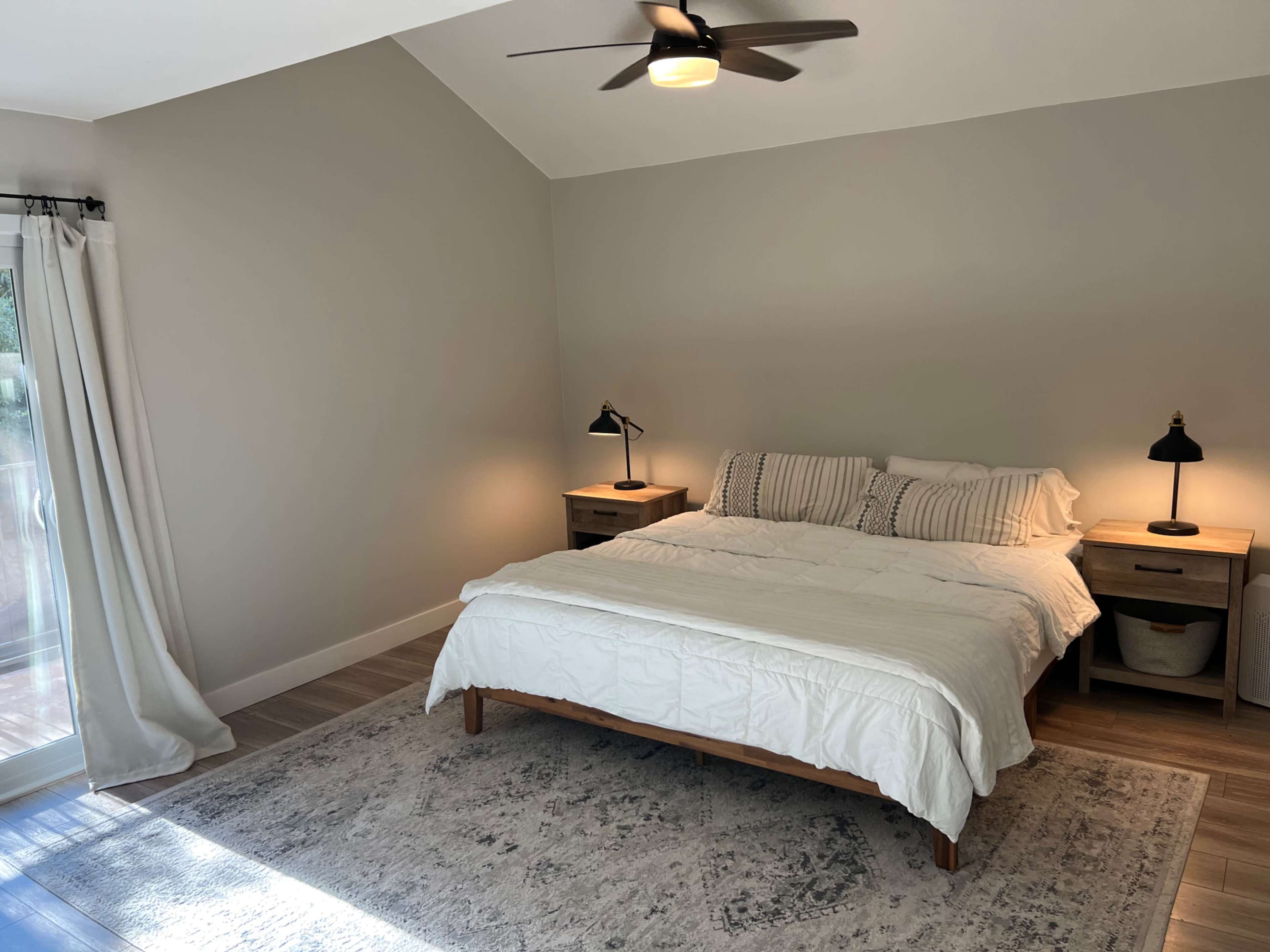 The image shows a minimalist bedroom with a bed, two bedside tables, and a large window allowing natural light.