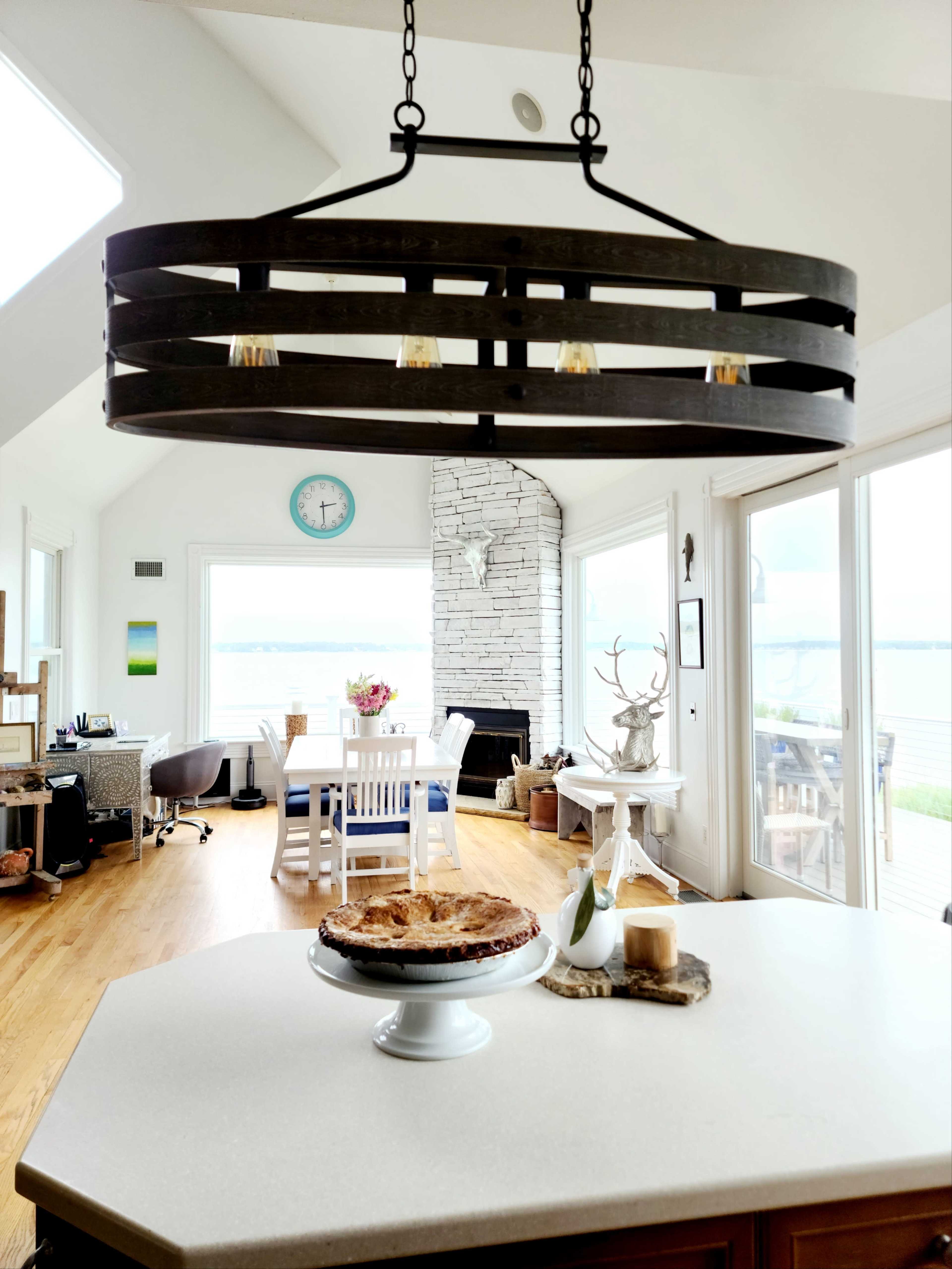 A spacious kitchen features a round wooden chandelier above a countertop, with a pie on display and a view of a dining area and large windows overlooking a body of water.