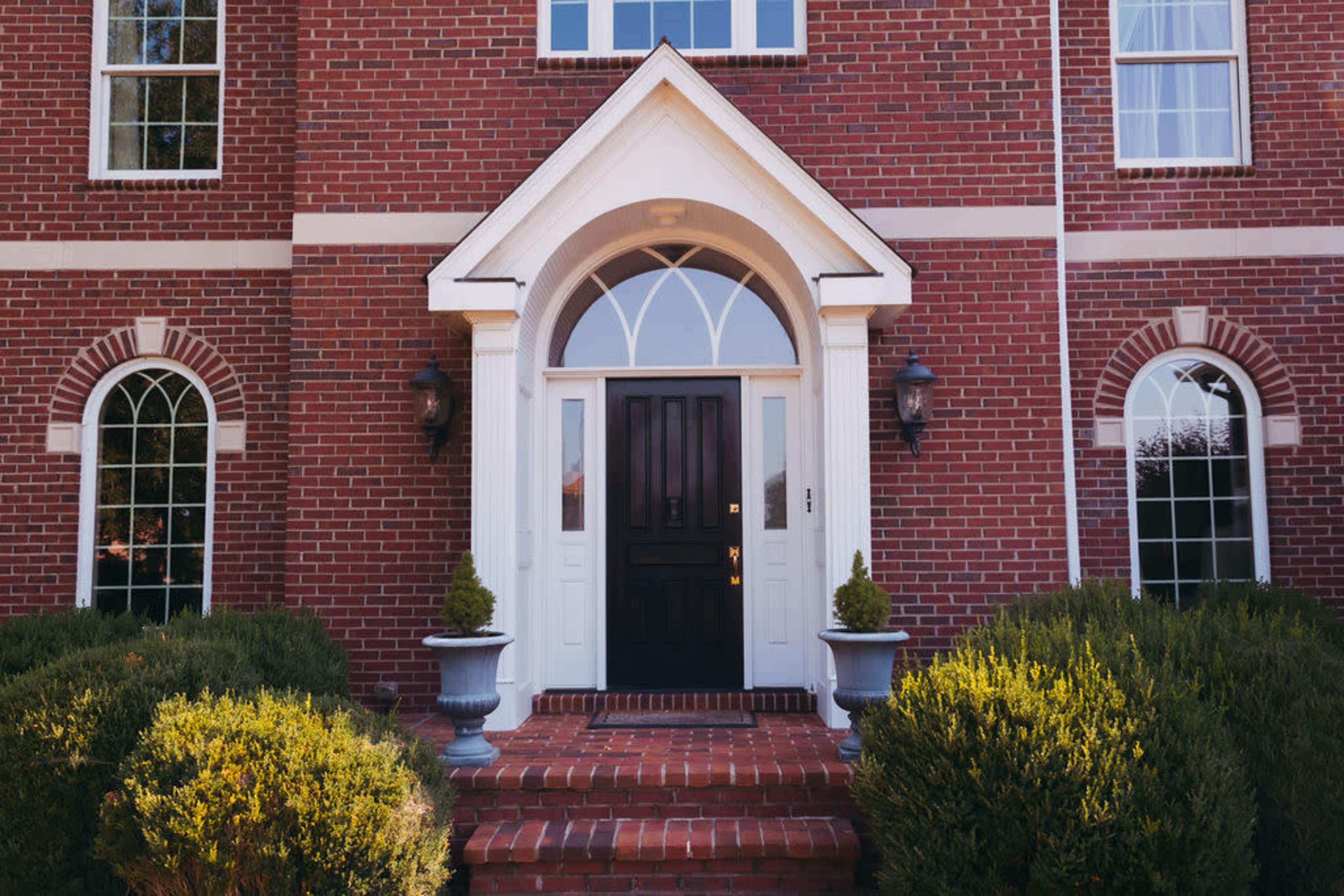 A brick house with a symmetrical front entrance featuring a black door, large windows on either side, and two potted plants on either side of the steps.