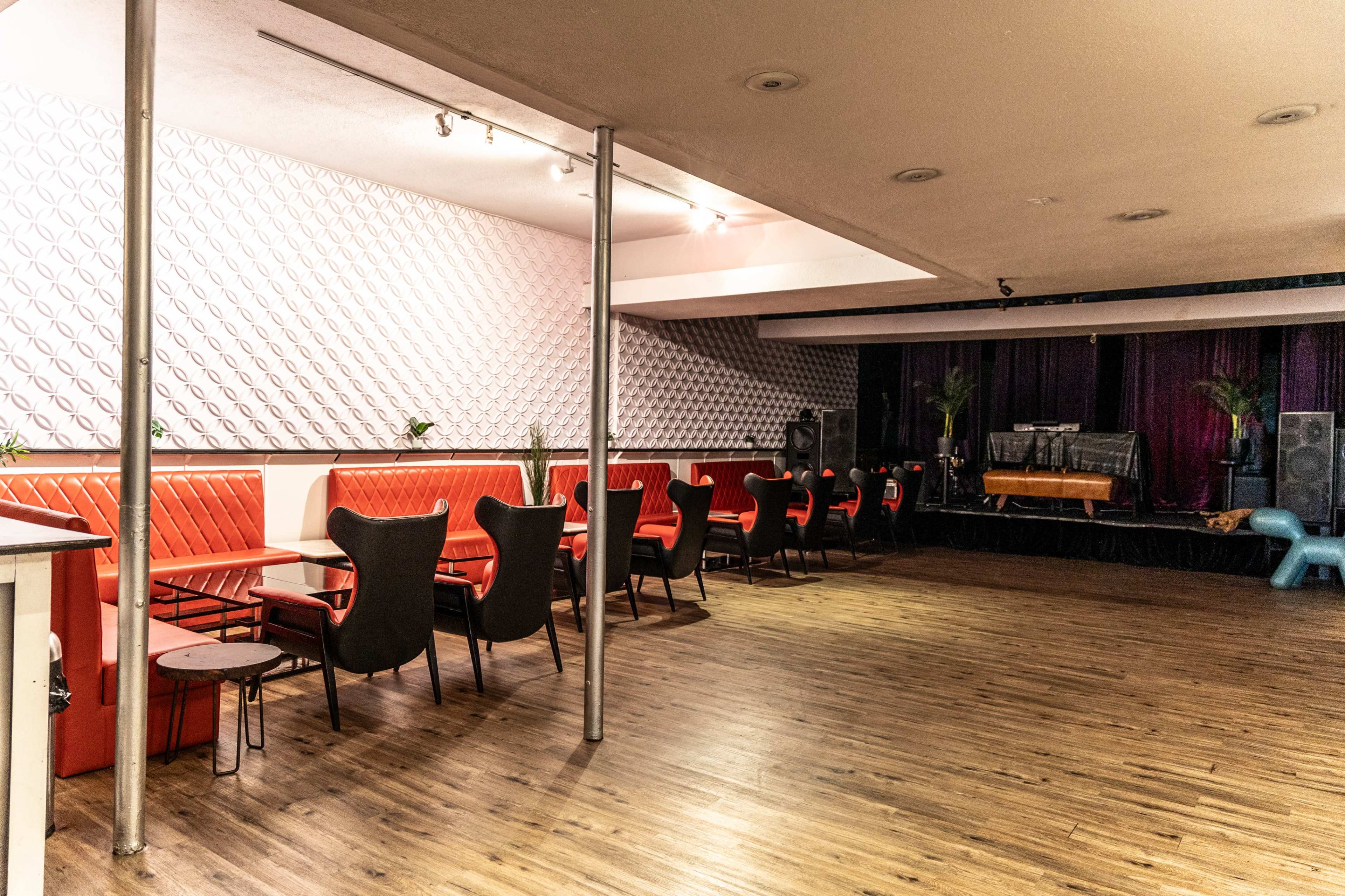 The image shows a spacious venue with red upholstered seating arranged around small tables, leading to a stage area with musical equipment in the background.
