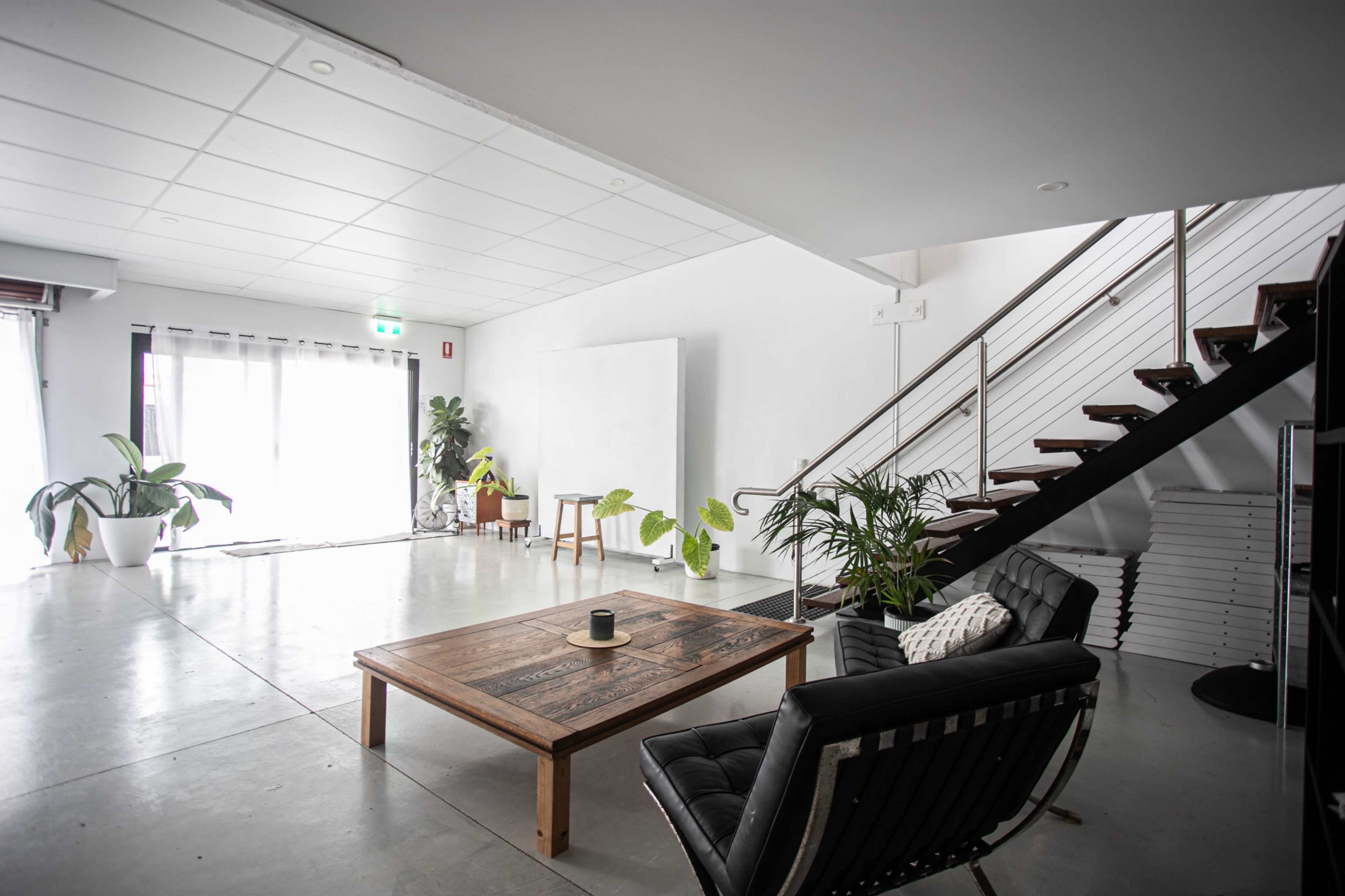 The image shows a modern, minimalistic living space featuring a wooden coffee table, two black chairs, a staircase, and several indoor plants.