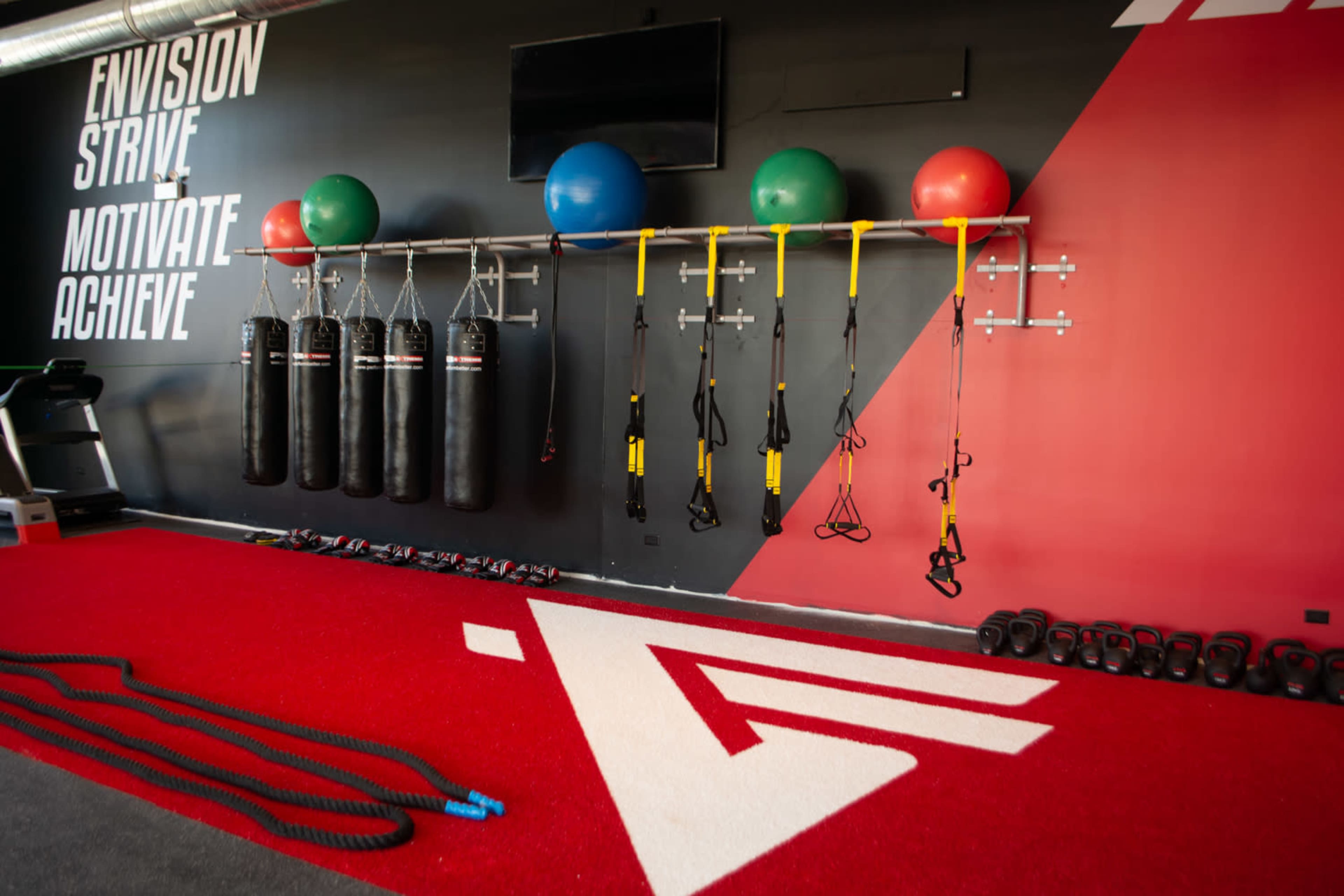 A fitness studio with a red and black wall featuring exercise equipment, including hanging punching bags, TRX straps, and stability balls.