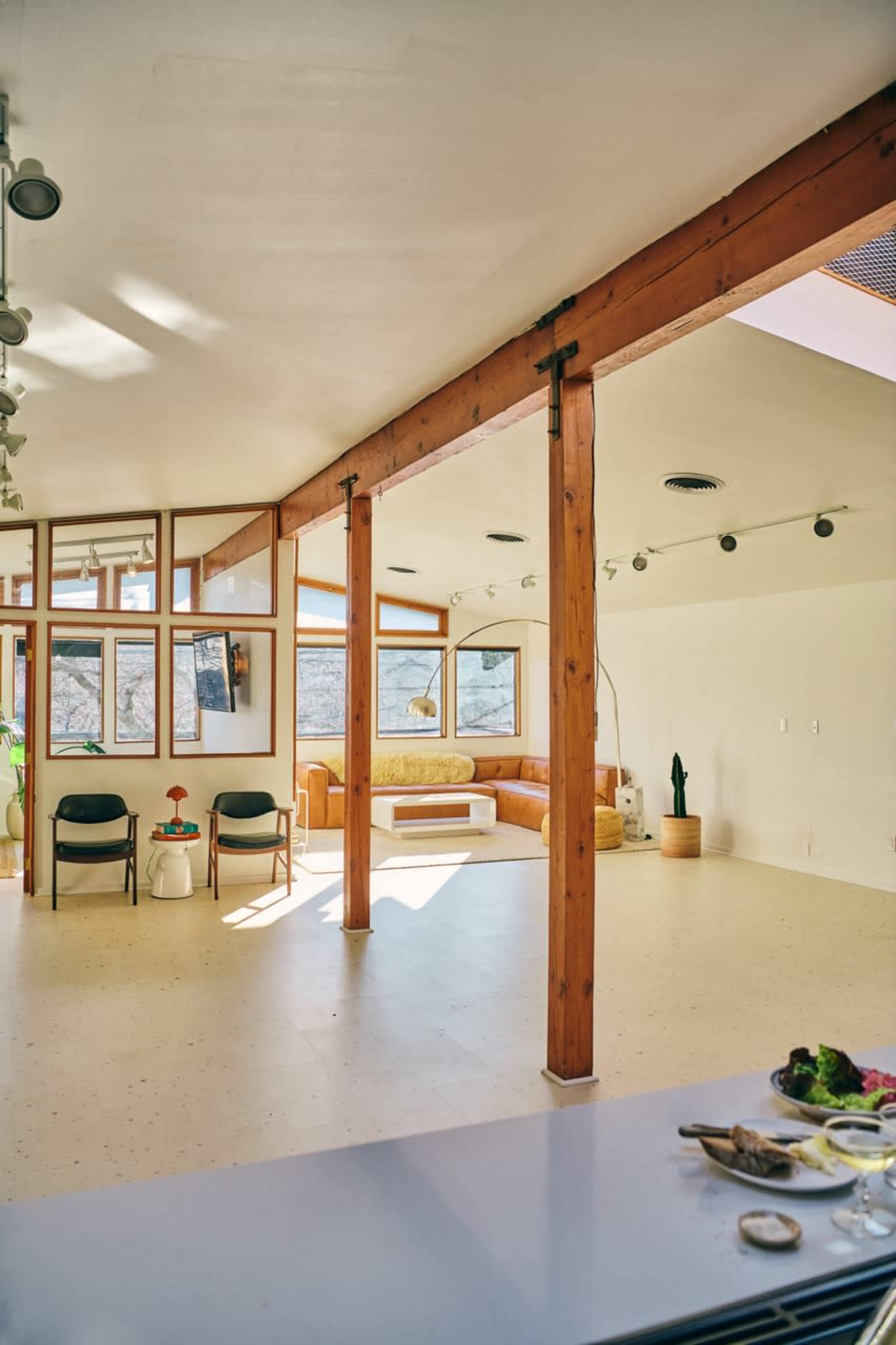 The image shows a spacious, modern interior featuring large windows, wooden pillars, and a cozy seating area, with a small table set in the foreground.