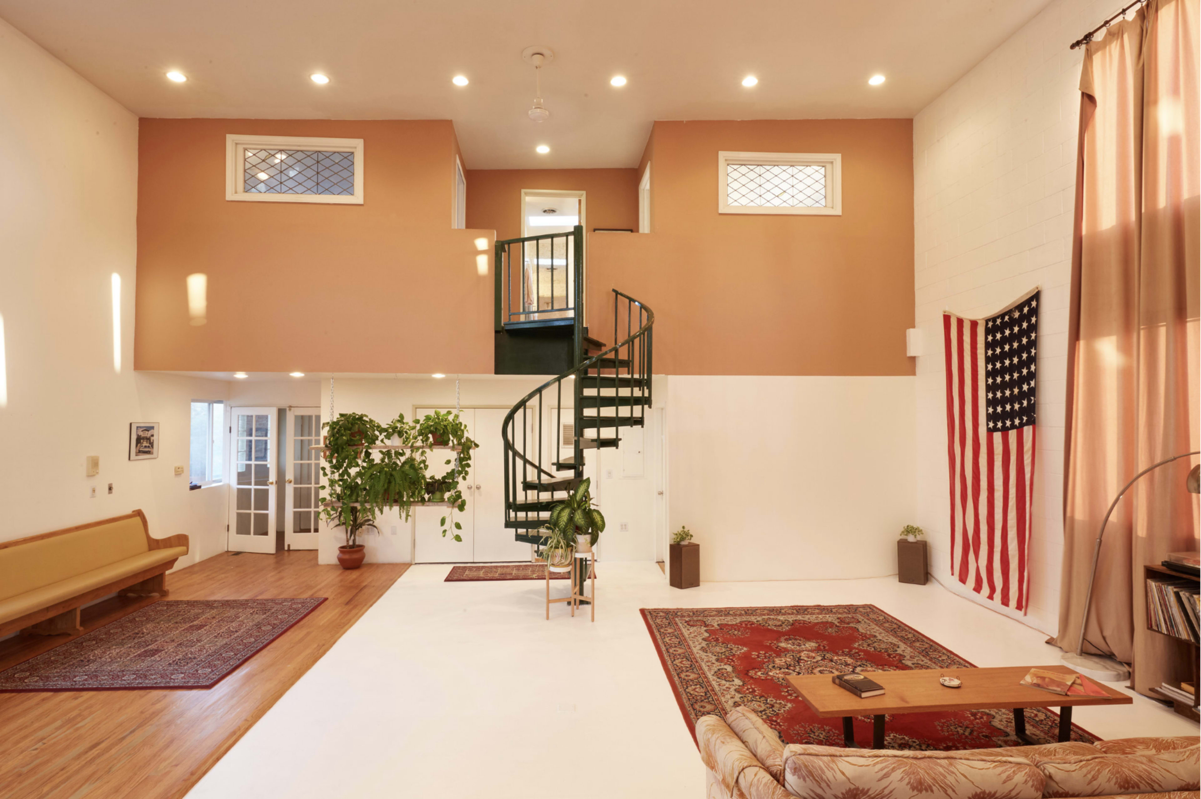 A spacious room features a spiral staircase, large windows, potted plants, and an American flag hanging on the wall, with rugs placed on a light-colored floor.