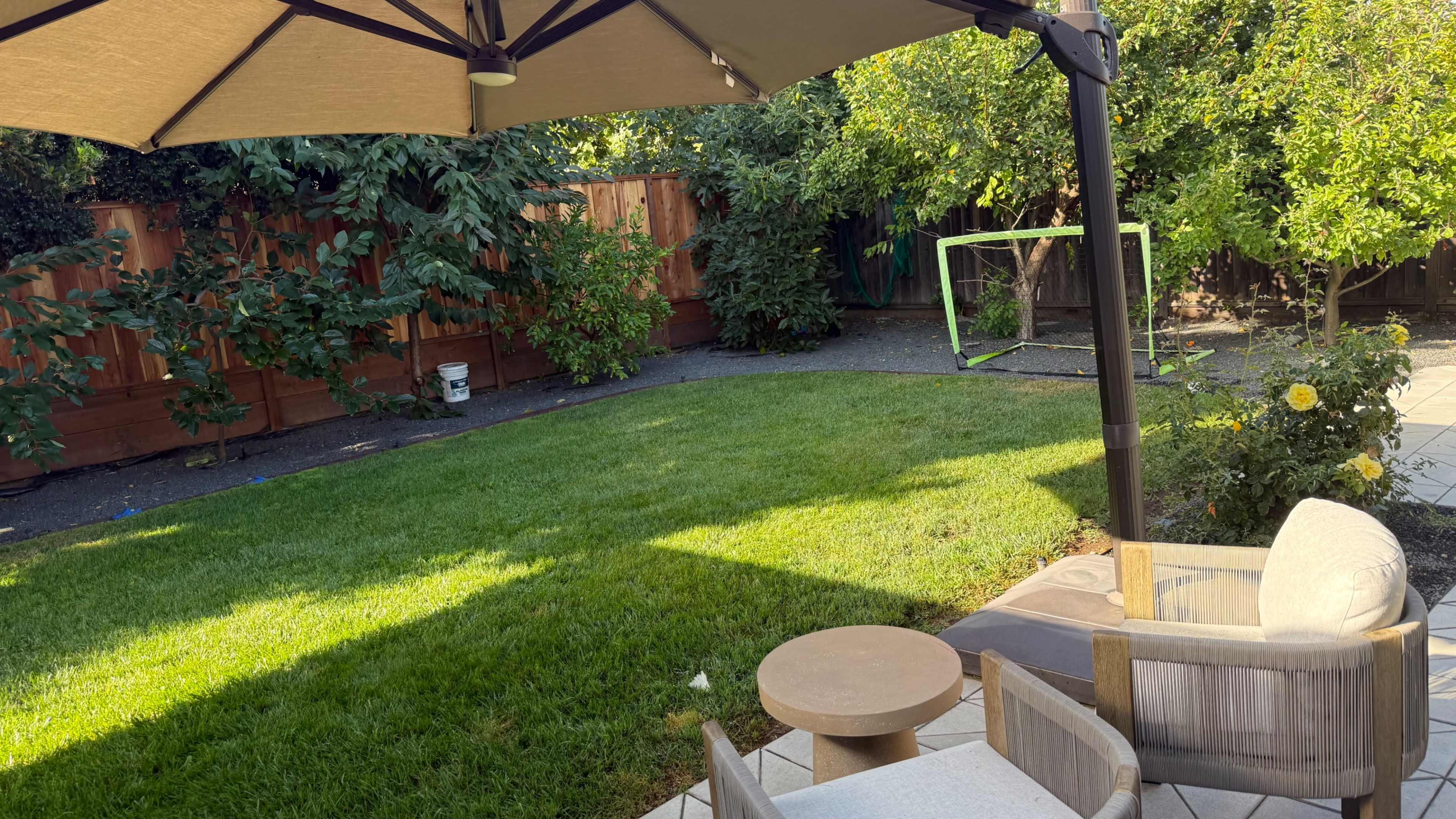 The image shows a backyard with a well-maintained lawn, a patio set, and a soccer goal in the distance, all shaded by a large umbrella.