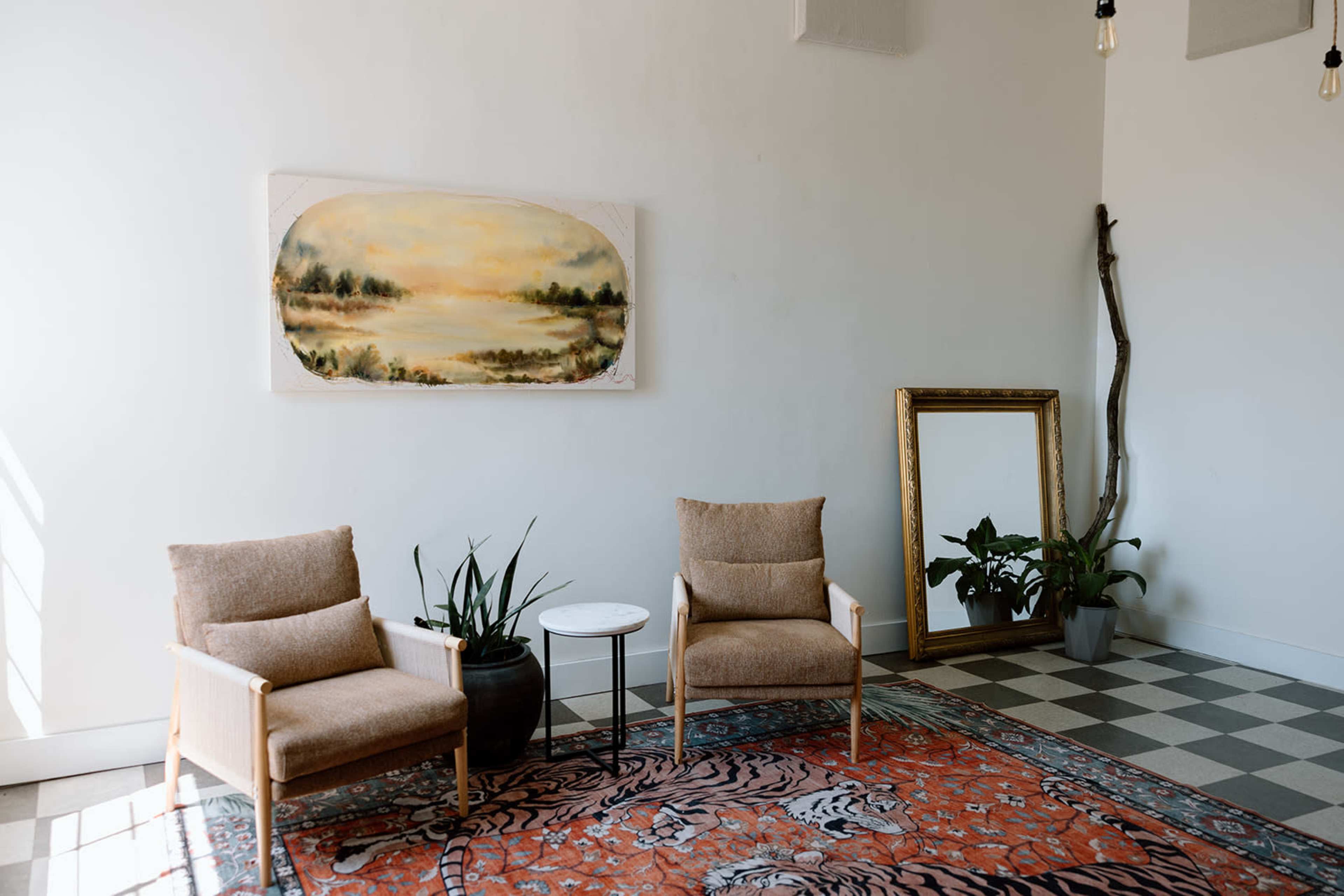 The image shows a bright room featuring two beige chairs, a small side table, a large mirror, an artwork of a landscape, and a potted plant on a patterned rug.