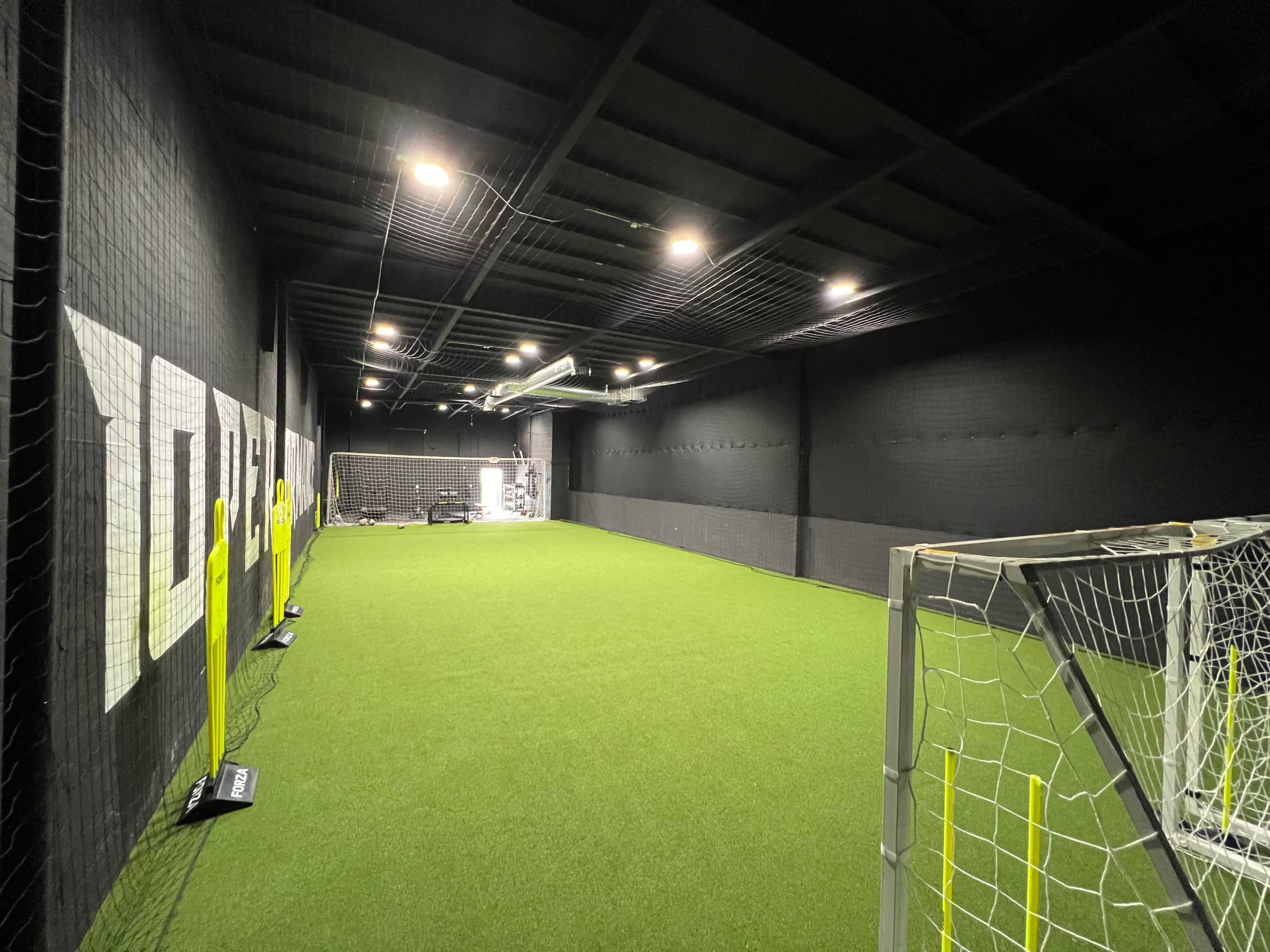 An indoor soccer training facility with artificial turf, two goalposts at either end, and black walls featuring large white letters.
