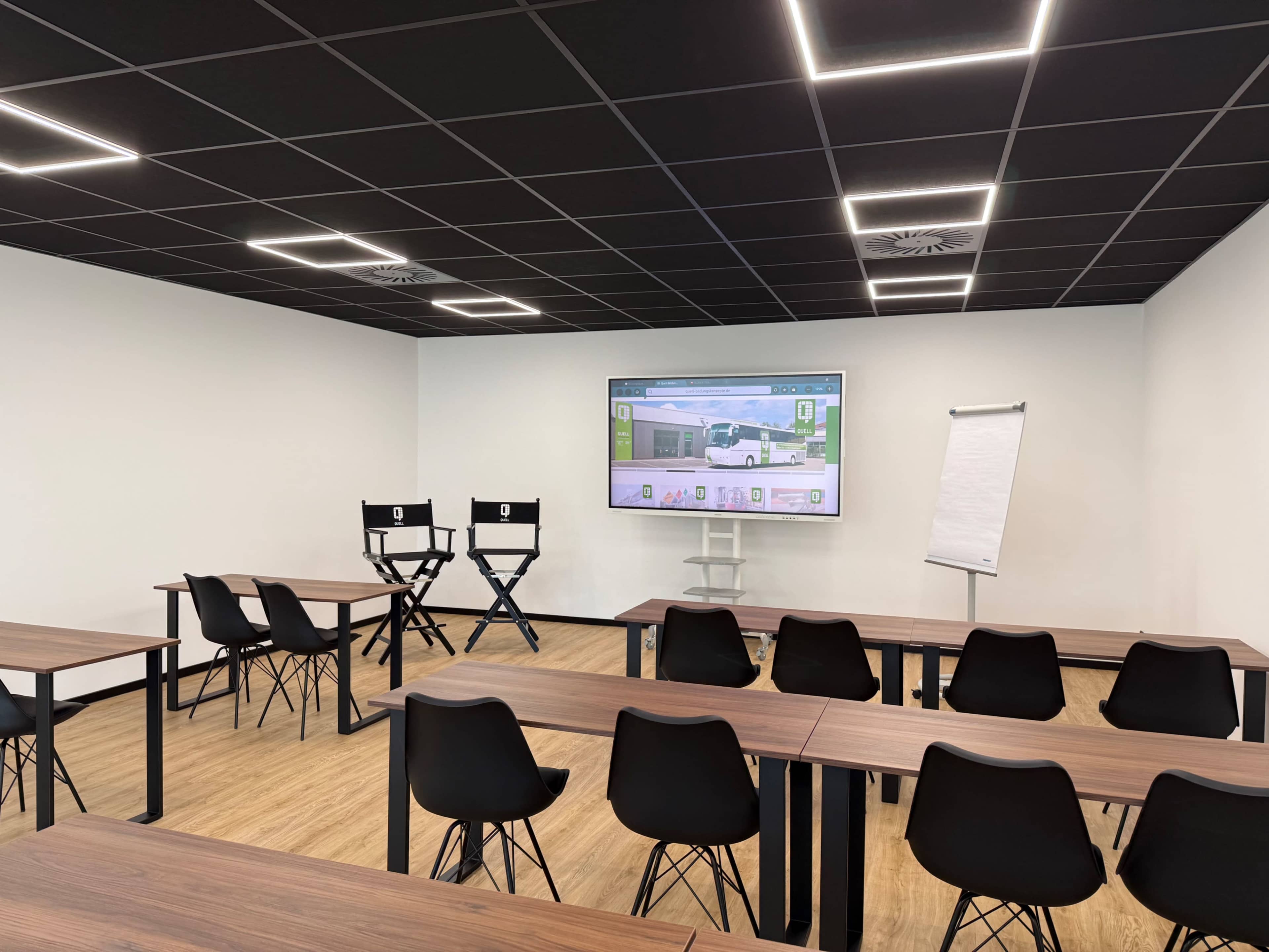 The image shows a modern classroom setup with rows of tables and chairs, a large screen displaying a presentation, and two director's chairs positioned nearby.