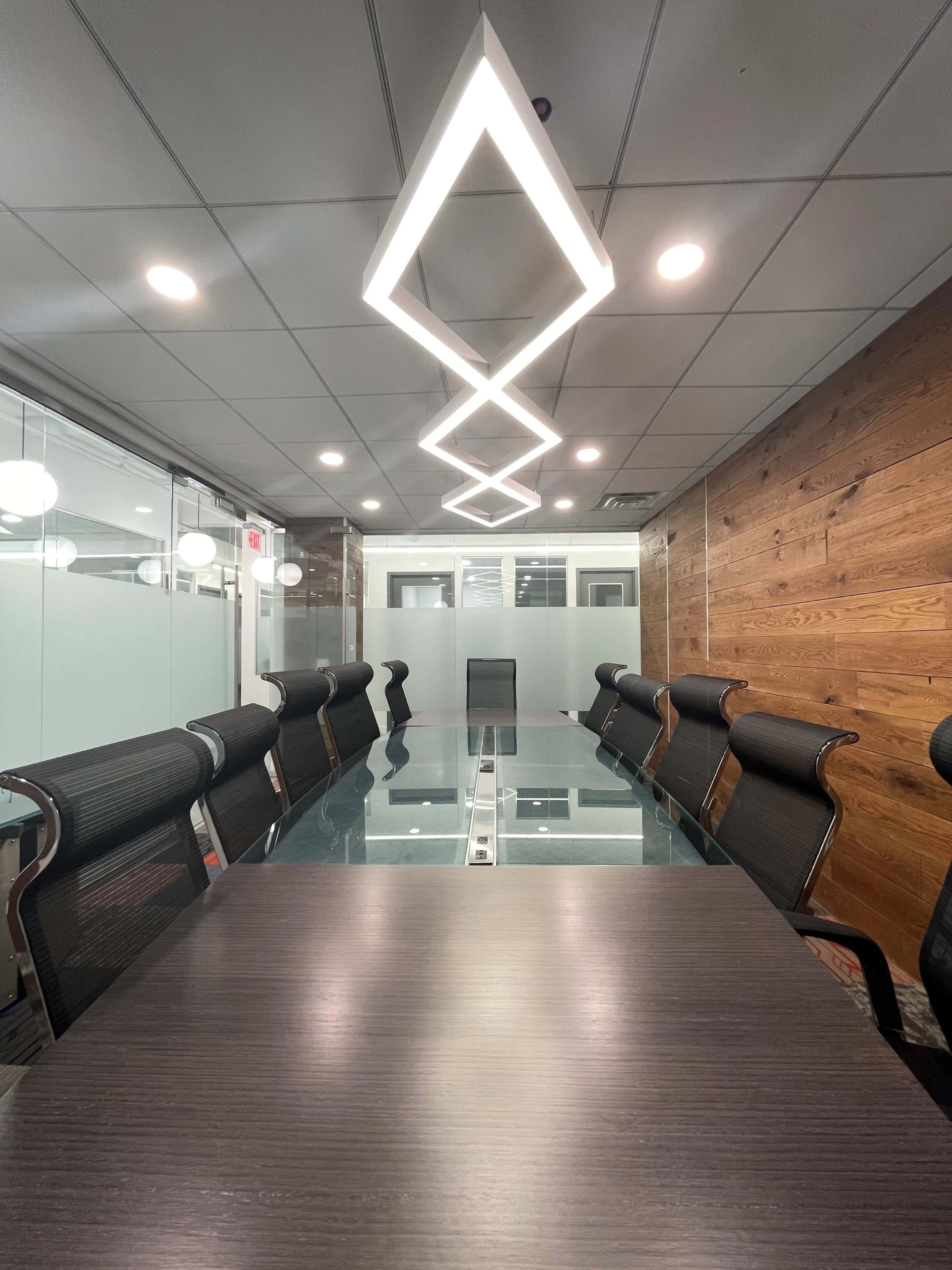 A modern conference room features a long glass table surrounded by ergonomic chairs, with geometric light fixtures above and wooden paneling on one wall.