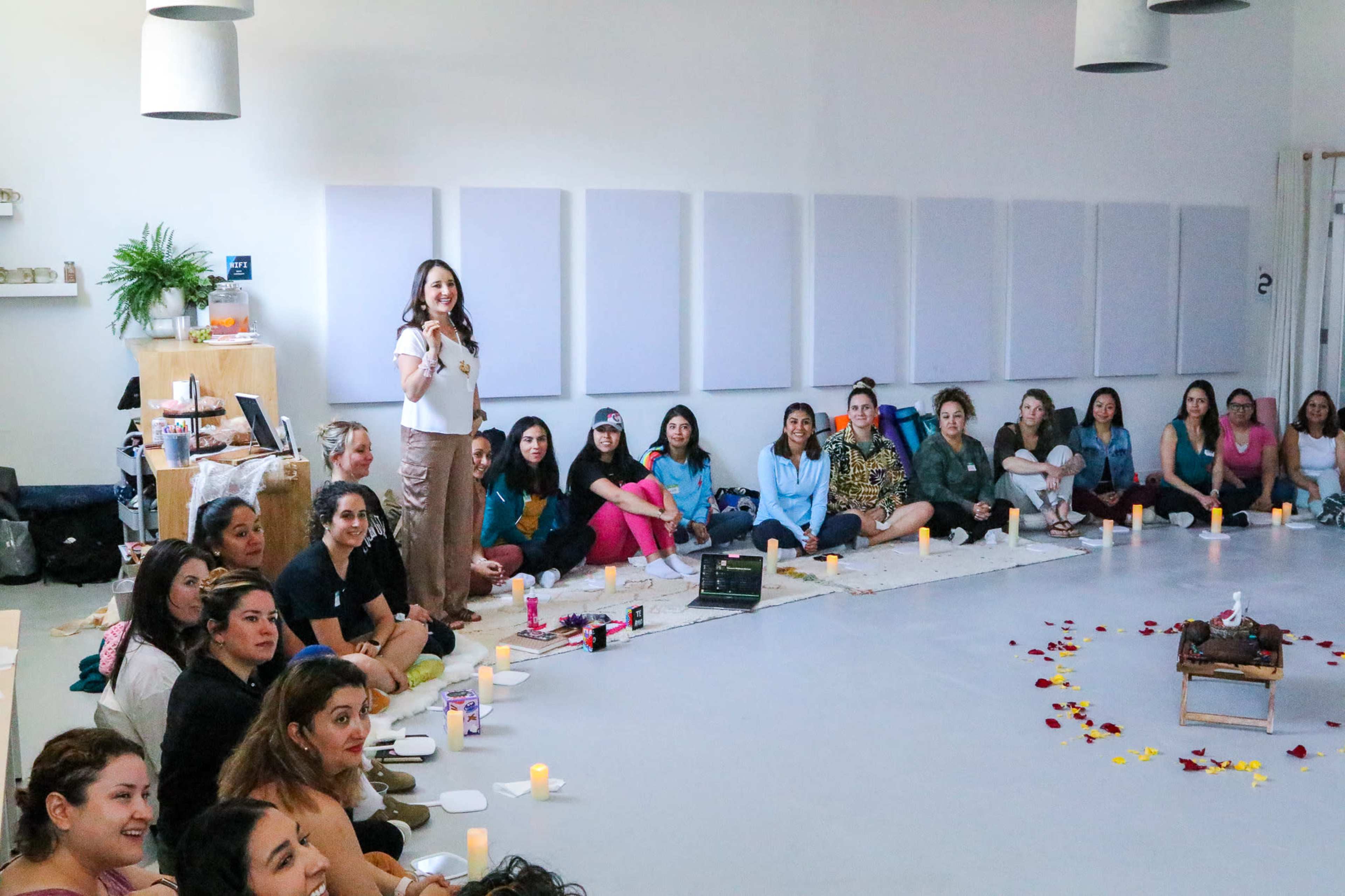 A group of women sits on the floor in a spacious room, attentively listening to a speaker while surrounded by candles and decorative petals.