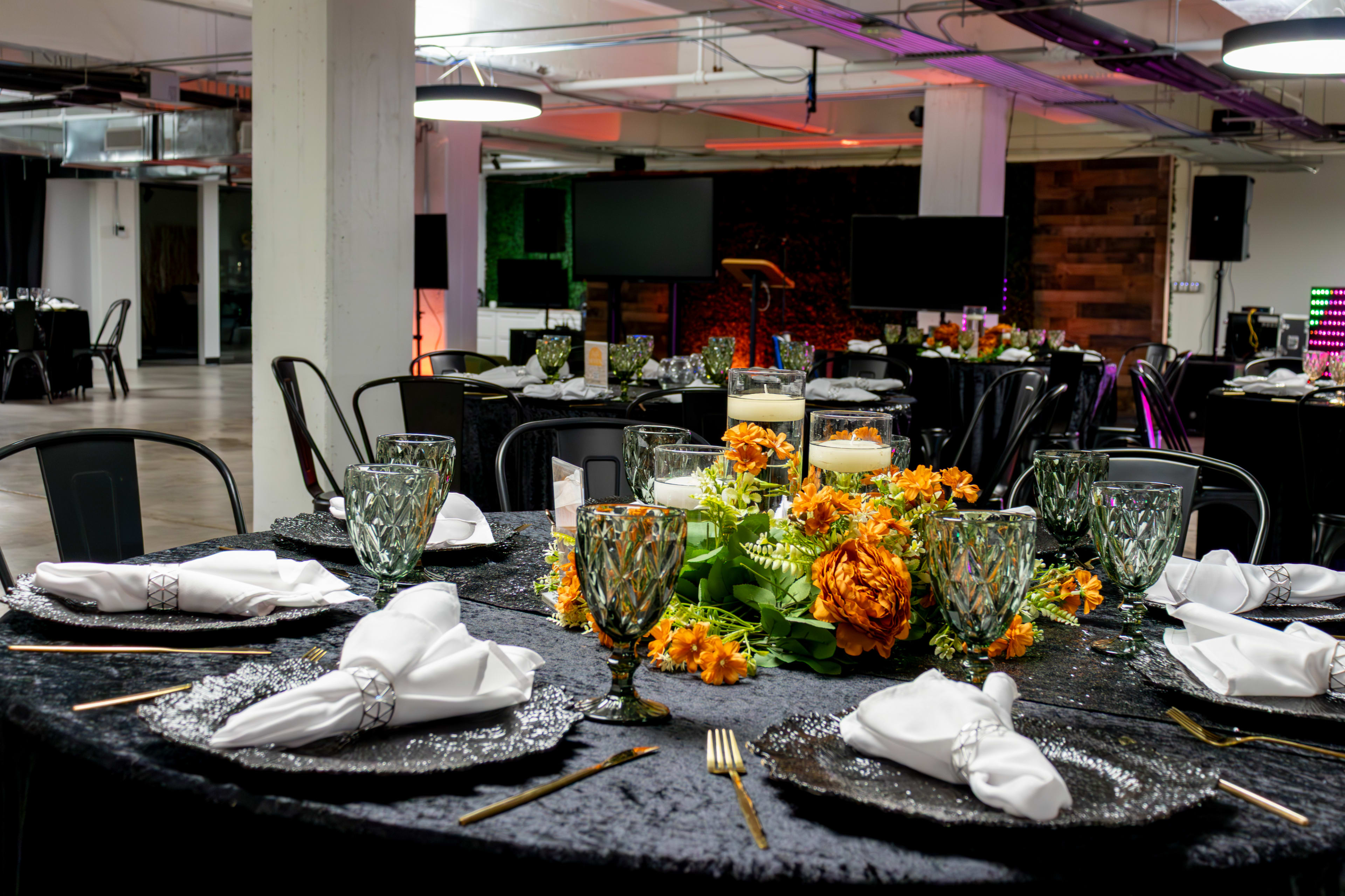 A dining setup features elegantly arranged tables with floral centerpieces, glassware, and neatly folded napkins in a modern event space.