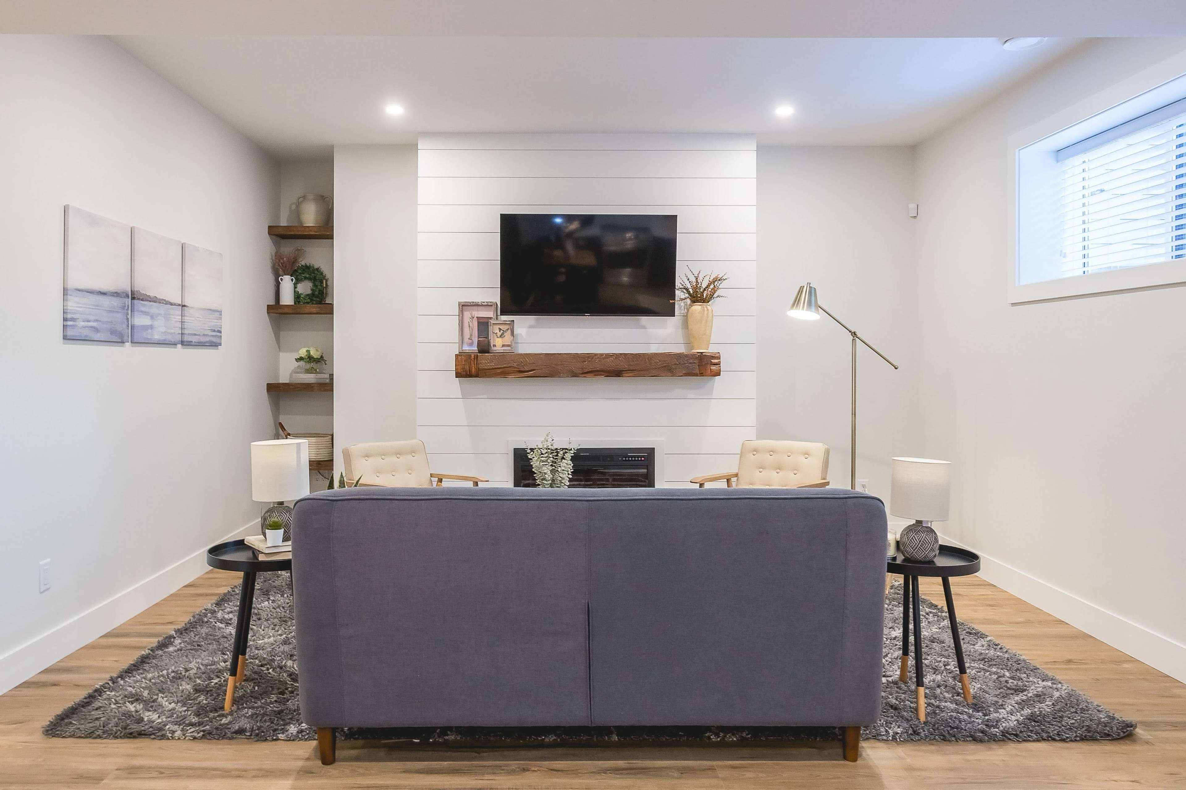 A modern living room features a gray sofa facing a wall-mounted television, flanked by two armchairs and lit by table lamps, with wooden shelves holding decorative items.