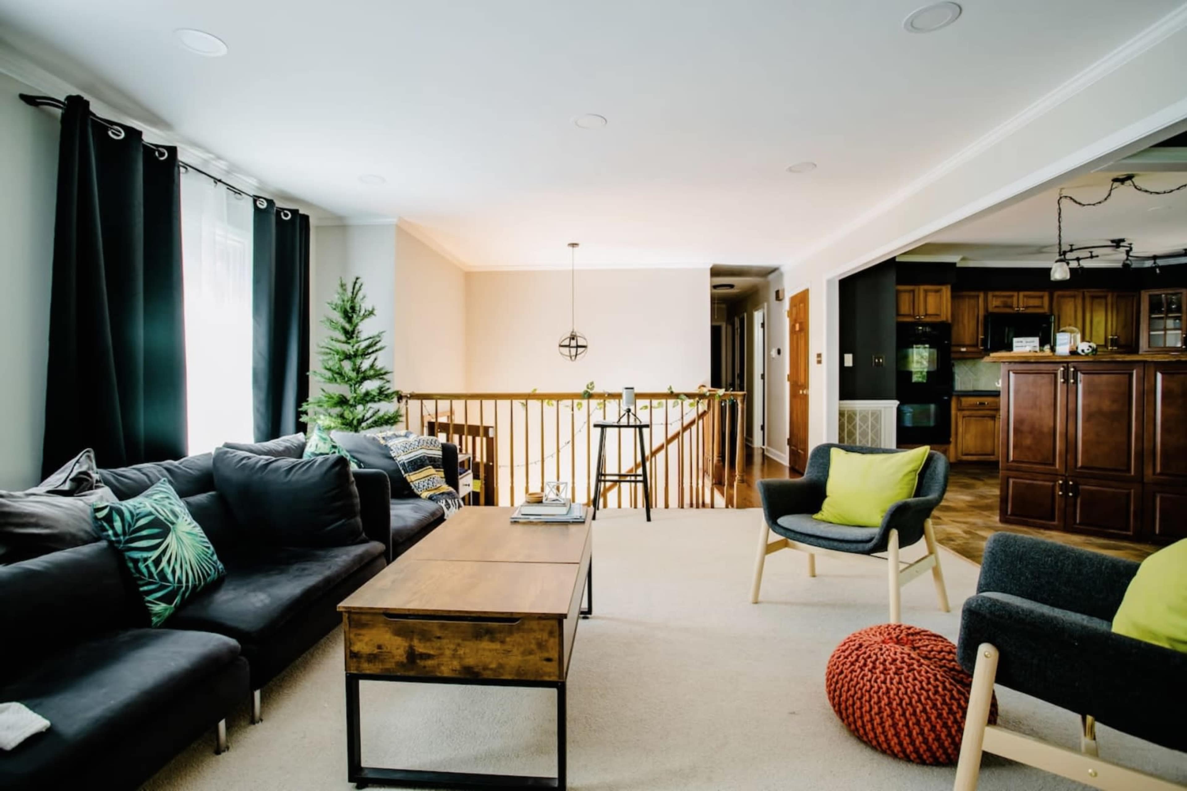 The image shows a living room with a black sectional sofa, a wooden coffee table, an armchair, and a decorative tree in the corner, featuring a staircase in the background.