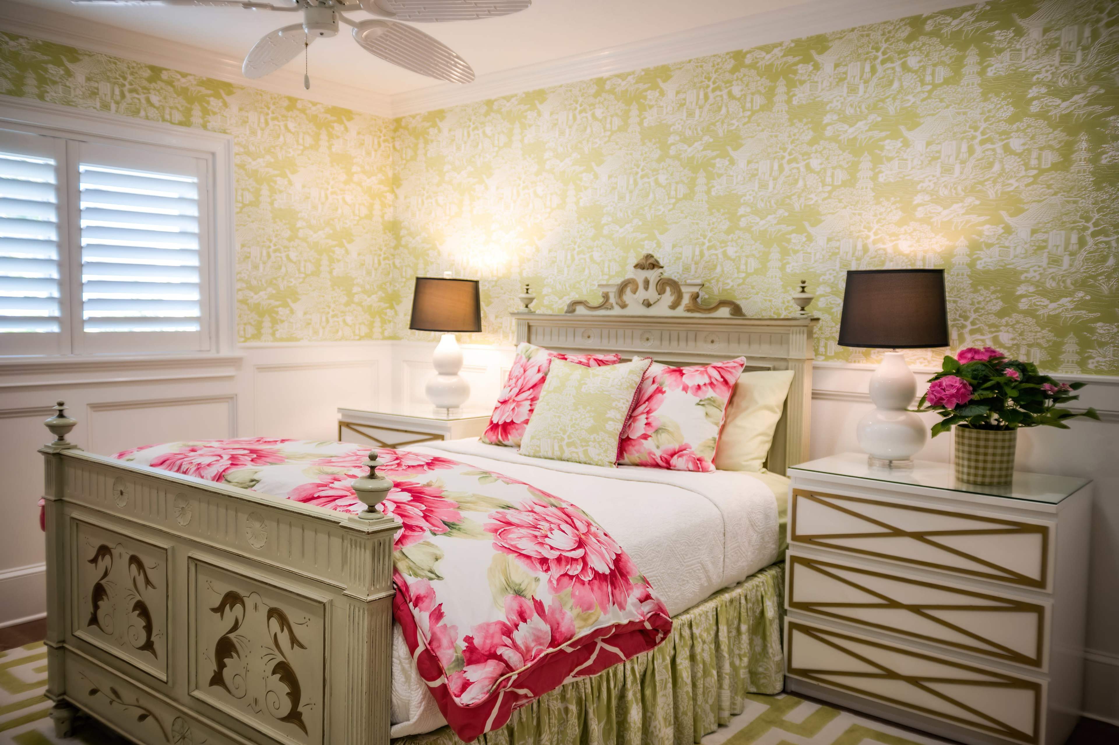 A bright bedroom featuring a white bed with floral bedding, a decorative headboard, two lamps on nightstands, and a green and white patterned wall.