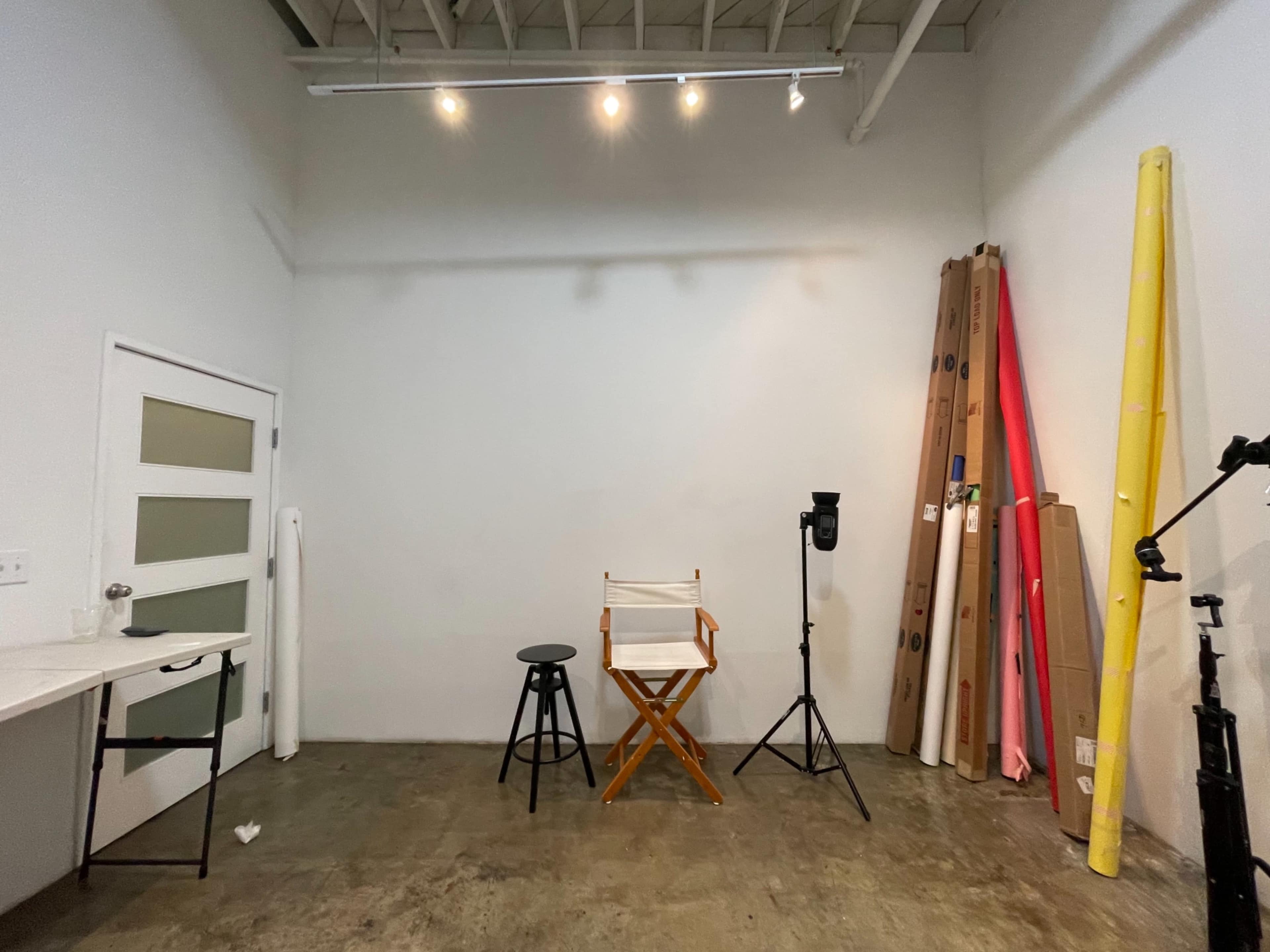 A photography studio features a director's chair, a stool, a backdrop stand, and rolls of colored paper against a white wall.