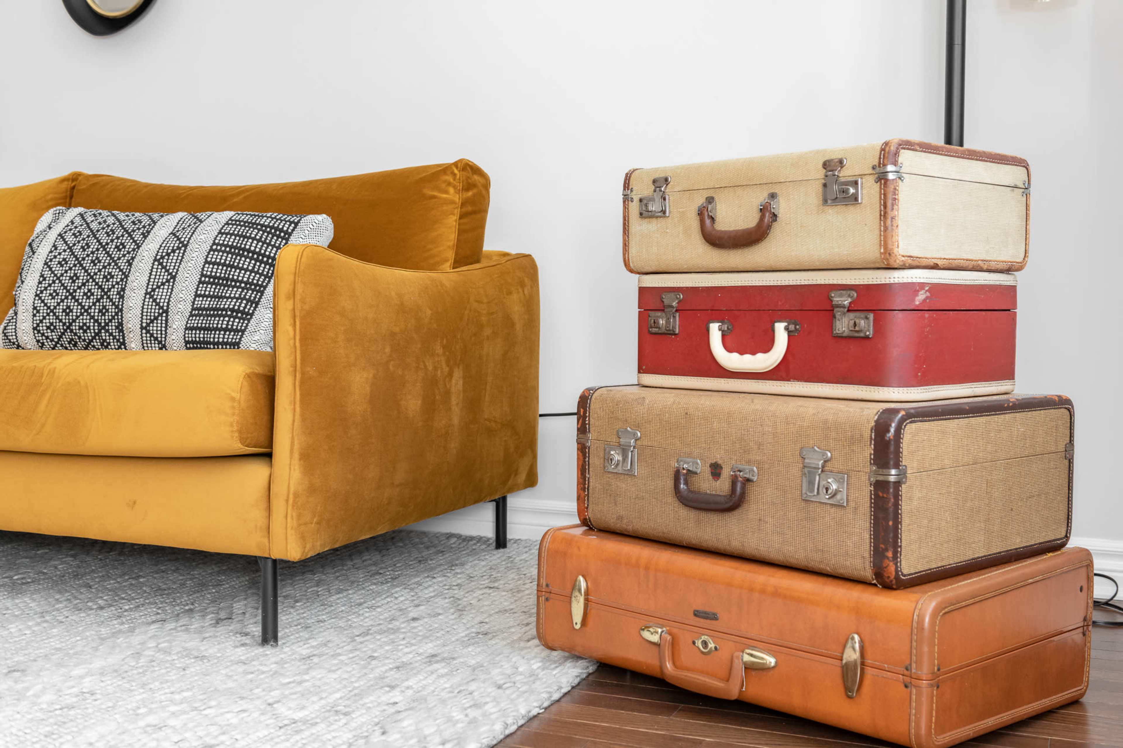 A stack of vintage suitcases is positioned beside a mustard-colored couch in a well-lit room.