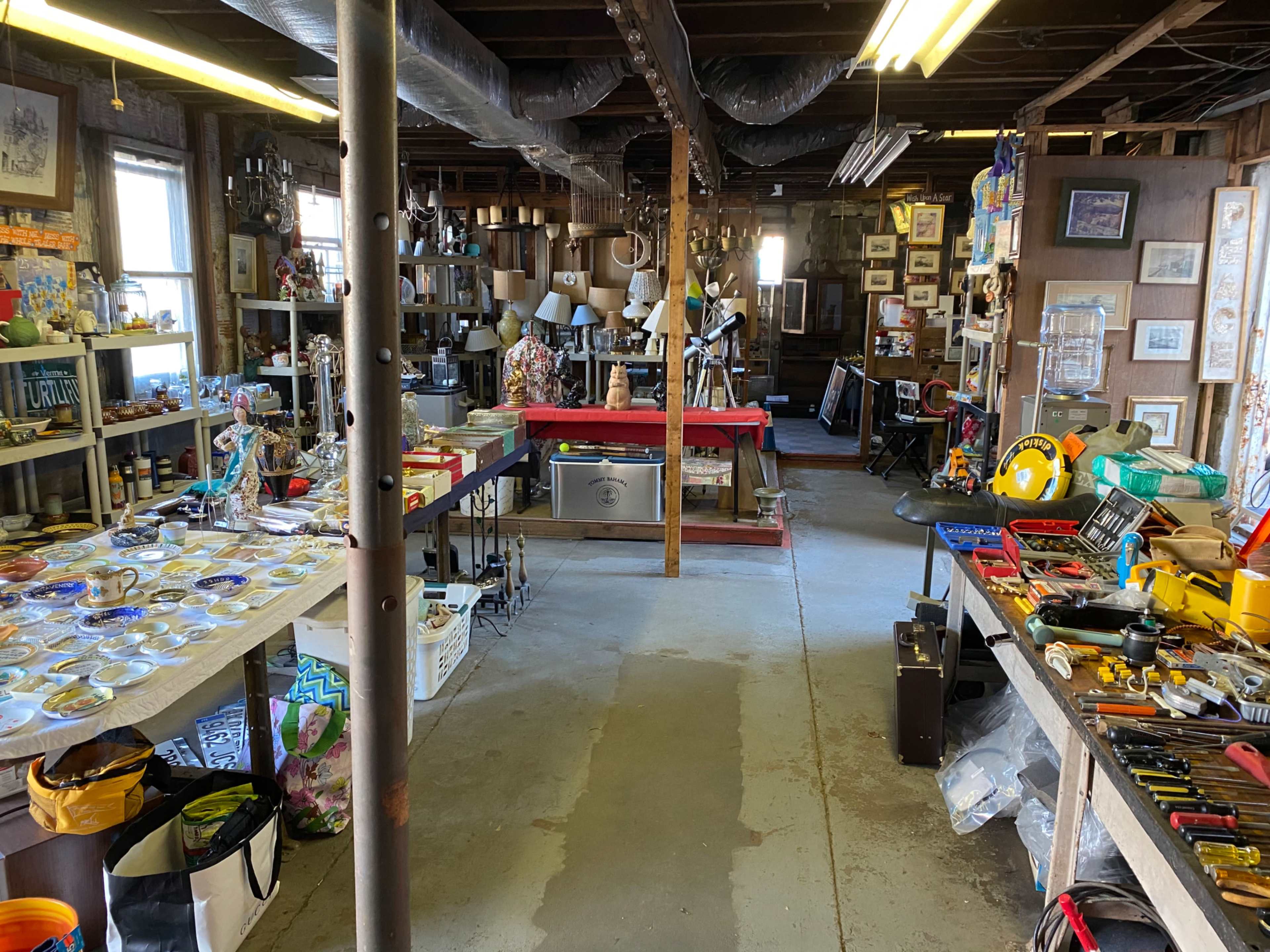 A large garage or workshop space filled with shelves of various tools, lamps, and decorative items, with tables displaying smaller objects on one side.