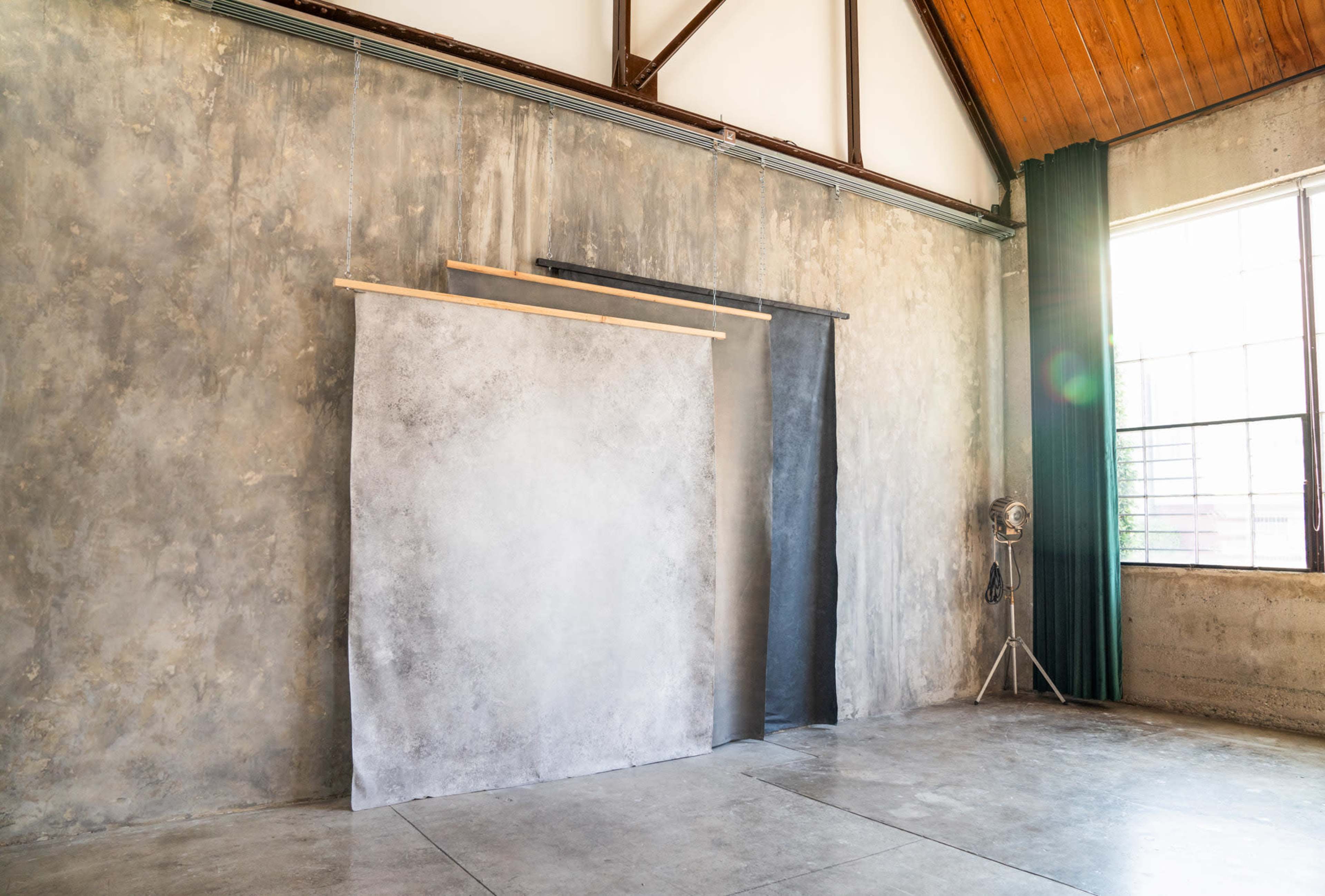 An industrial-style studio with a concrete wall, wooden beams overhead, and three backdrop panels hanging near a large window.