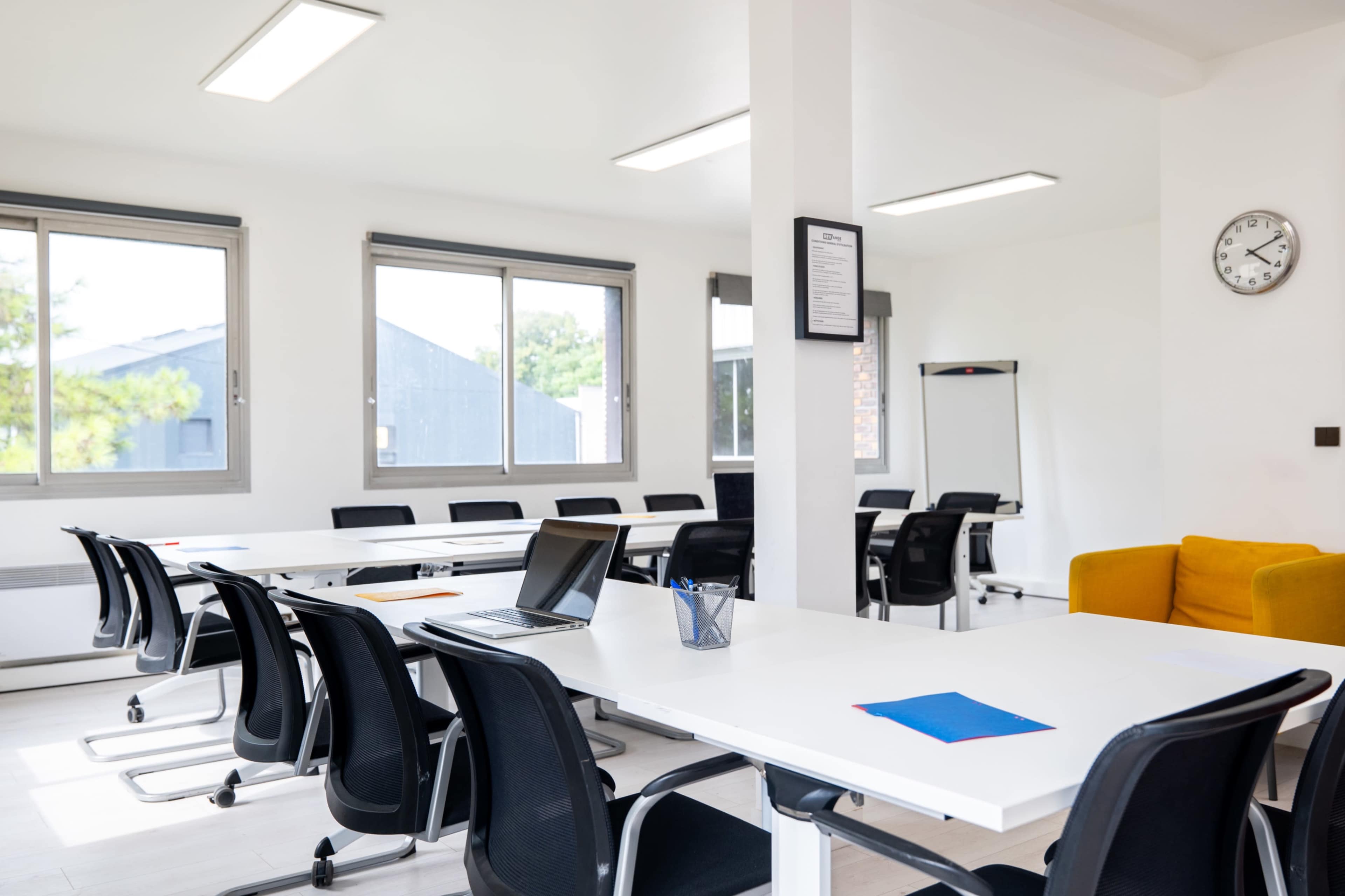 A modern meeting room features a long table with black chairs, a laptop, and colorful stationery, along with large windows allowing natural light.