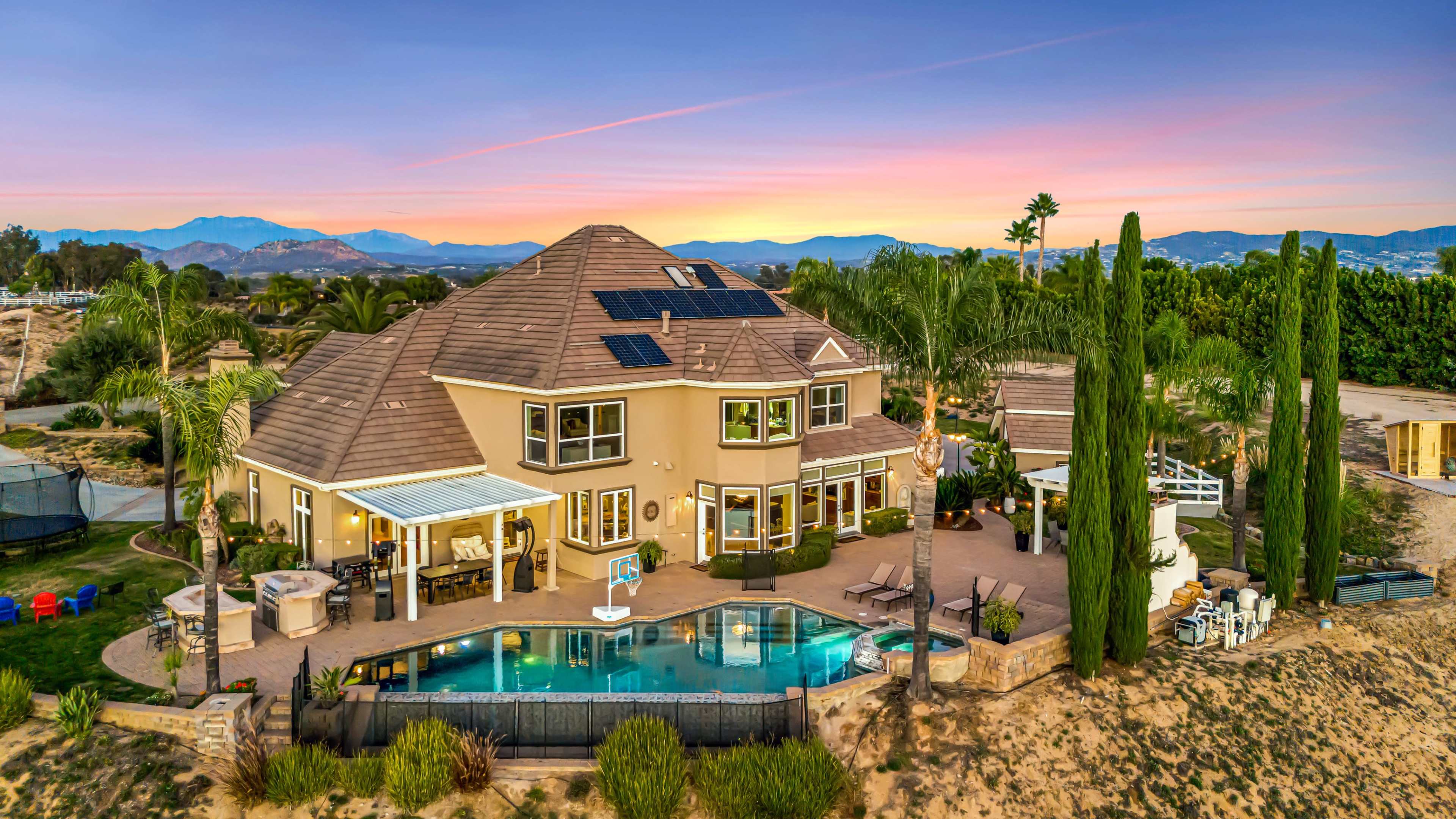 A large two-story house with a swimming pool and palm trees is set against a colorful sunset backdrop and mountains in the distance.