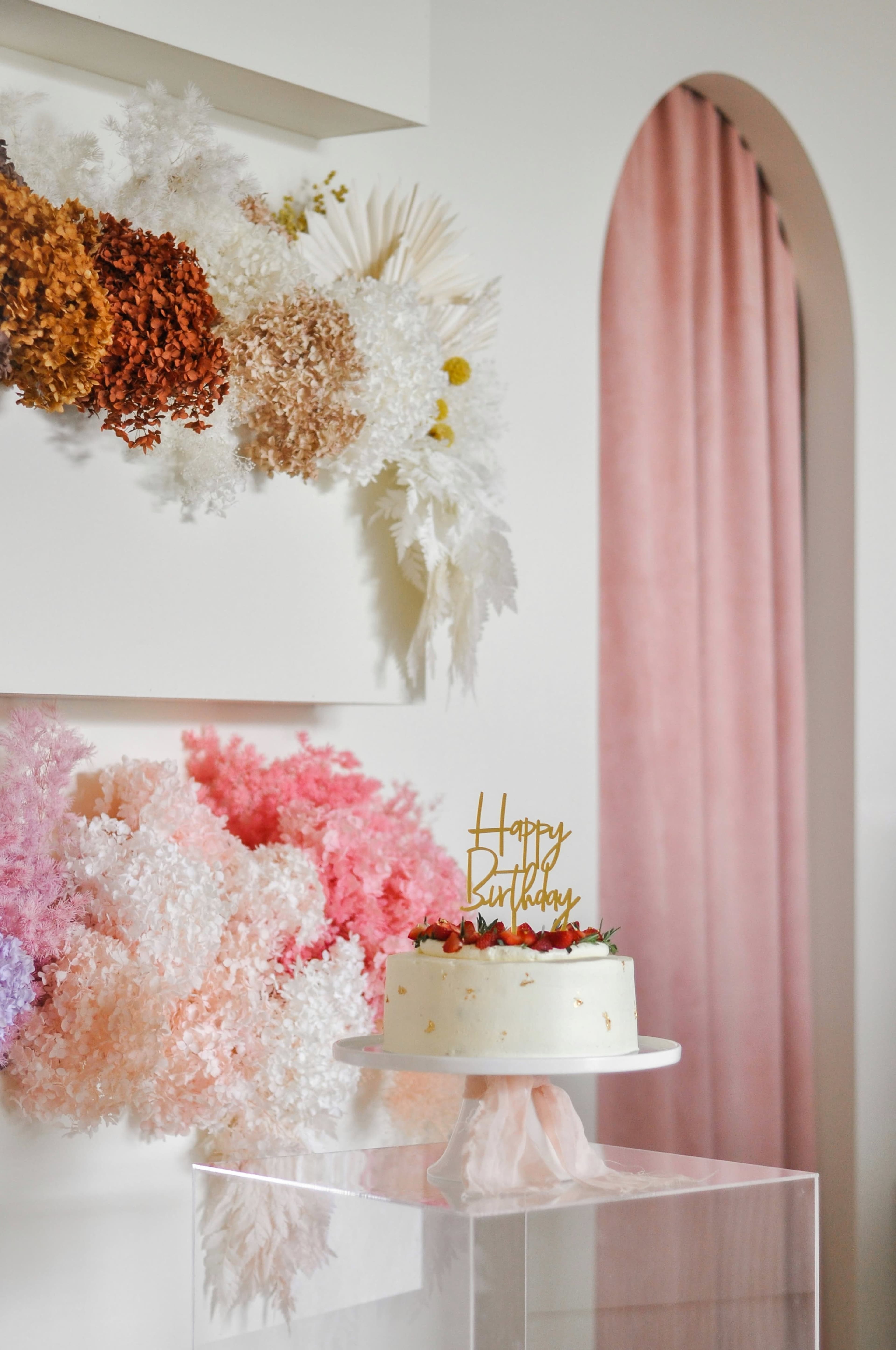 A white birthday cake with a "Happy Birthday" topper is displayed on a pedestal in front of a colorful floral arrangement against a stylish backdrop.