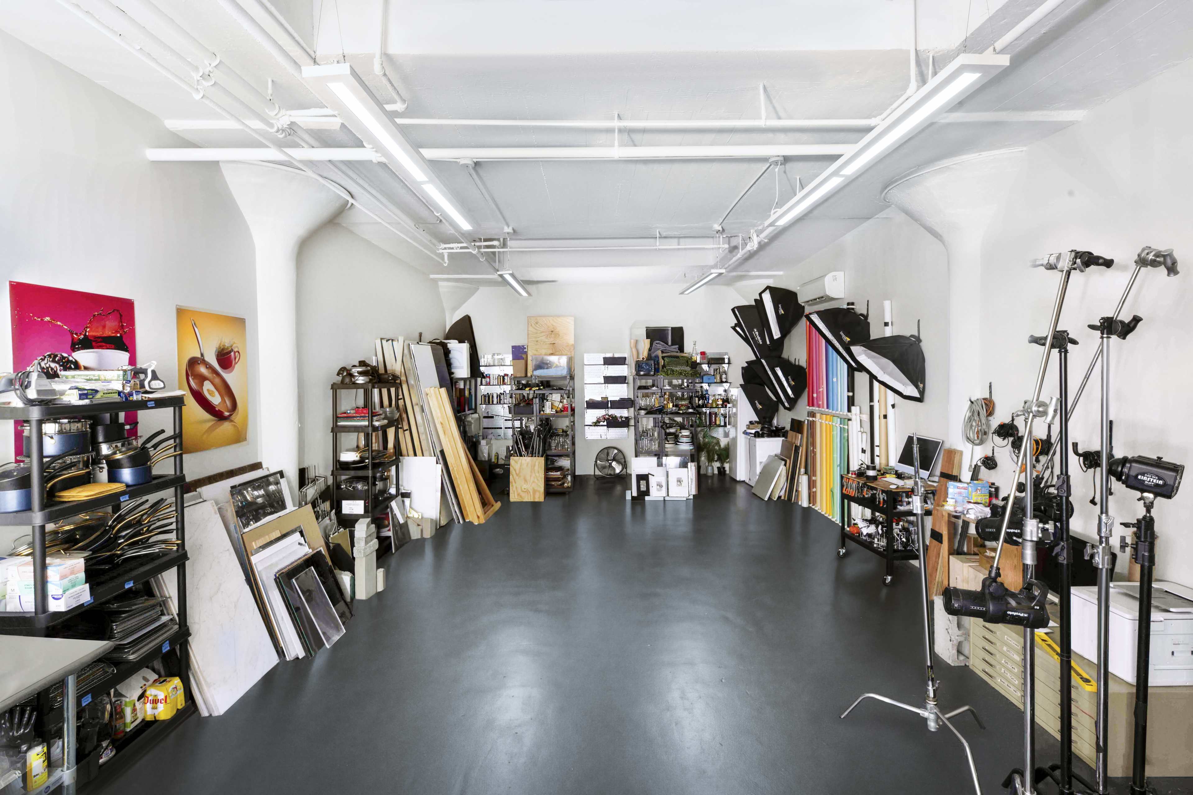 The image shows a spacious studio filled with various equipment, shelves, and materials organized along the walls.
