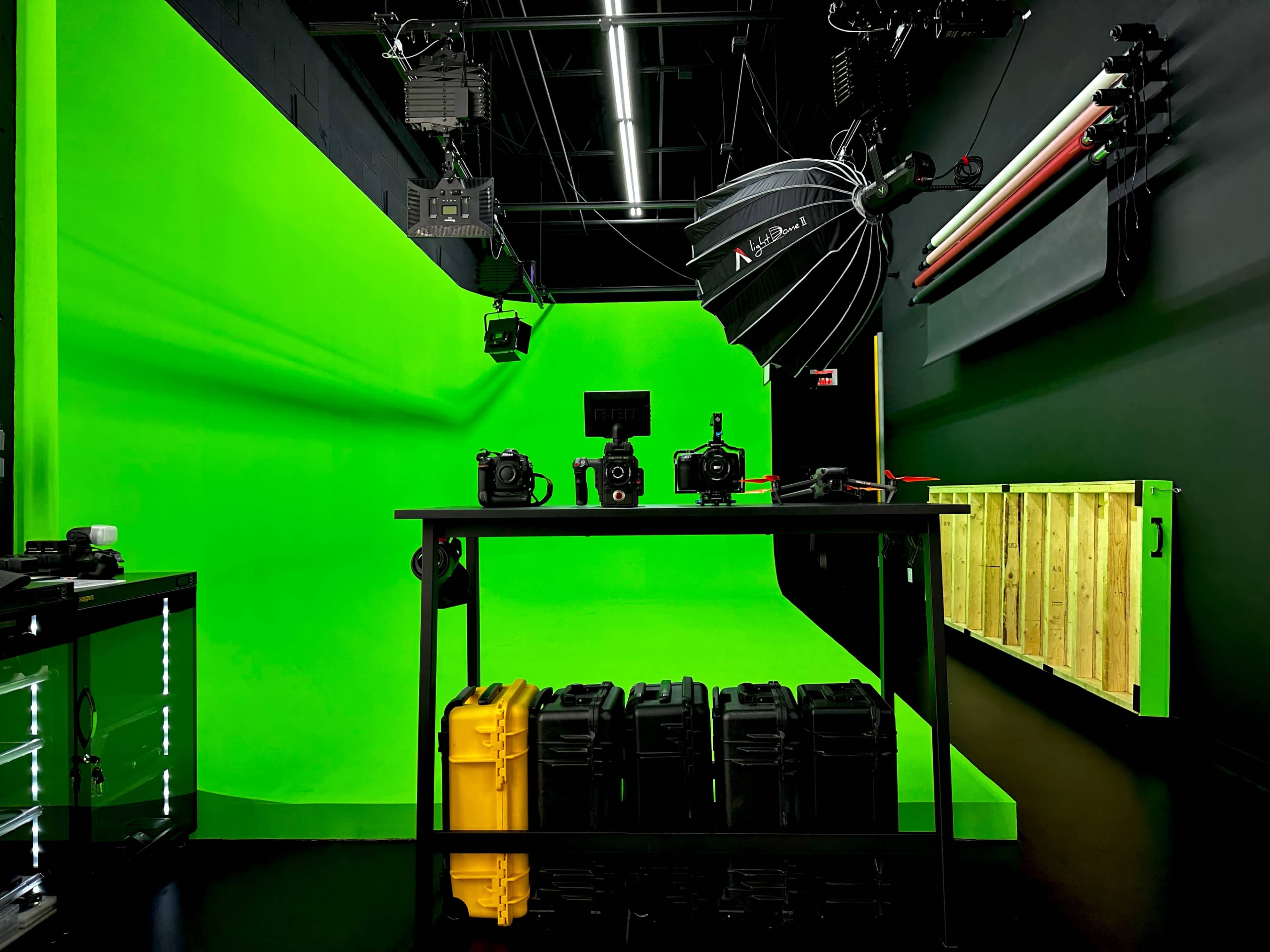 The image shows a photography studio with a green screen background, a black table holding several cameras, and various lighting equipment mounted on the ceiling.