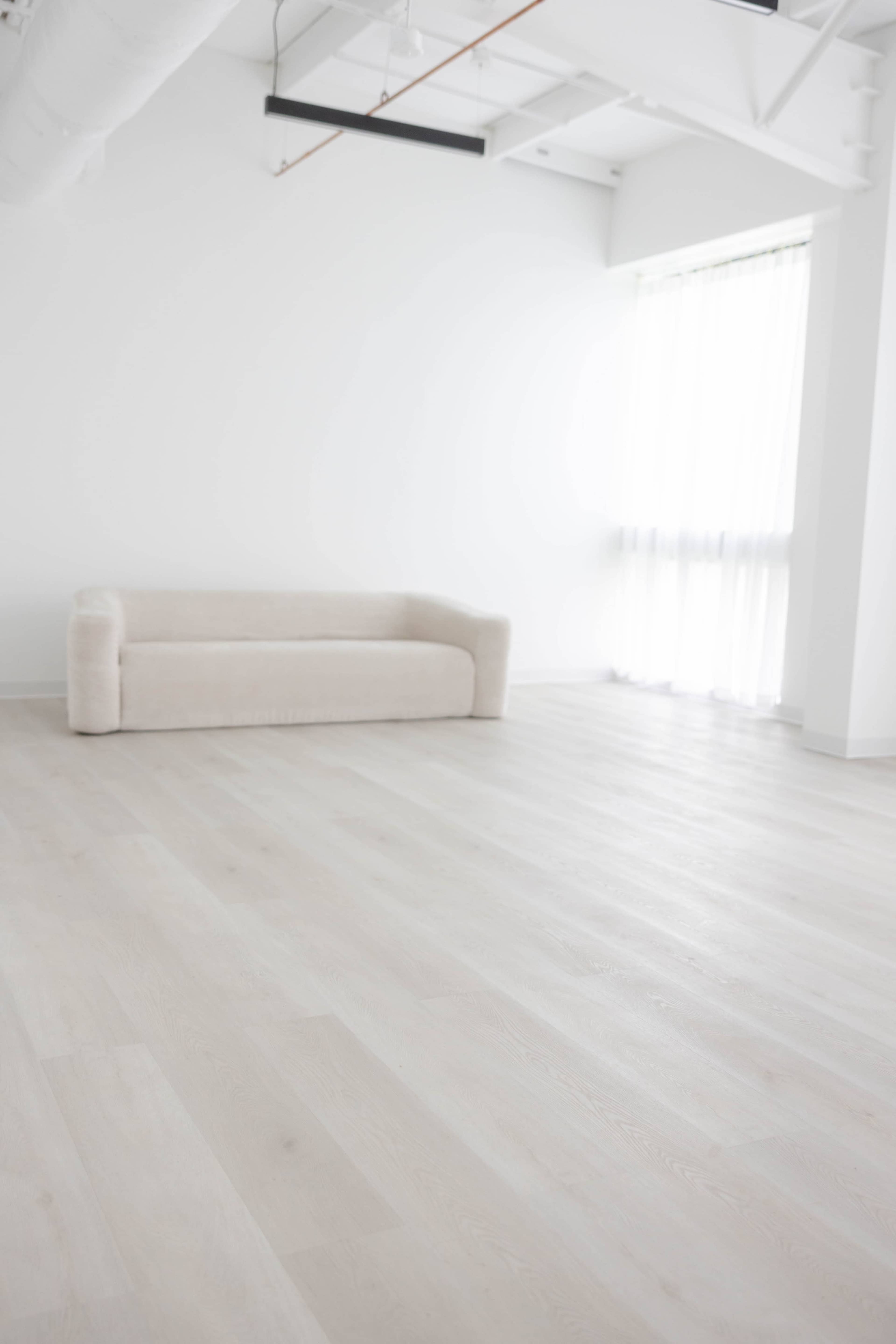 A light-filled, minimalist room features a beige sofa against a white wall, with large windows allowing natural light to stream in.