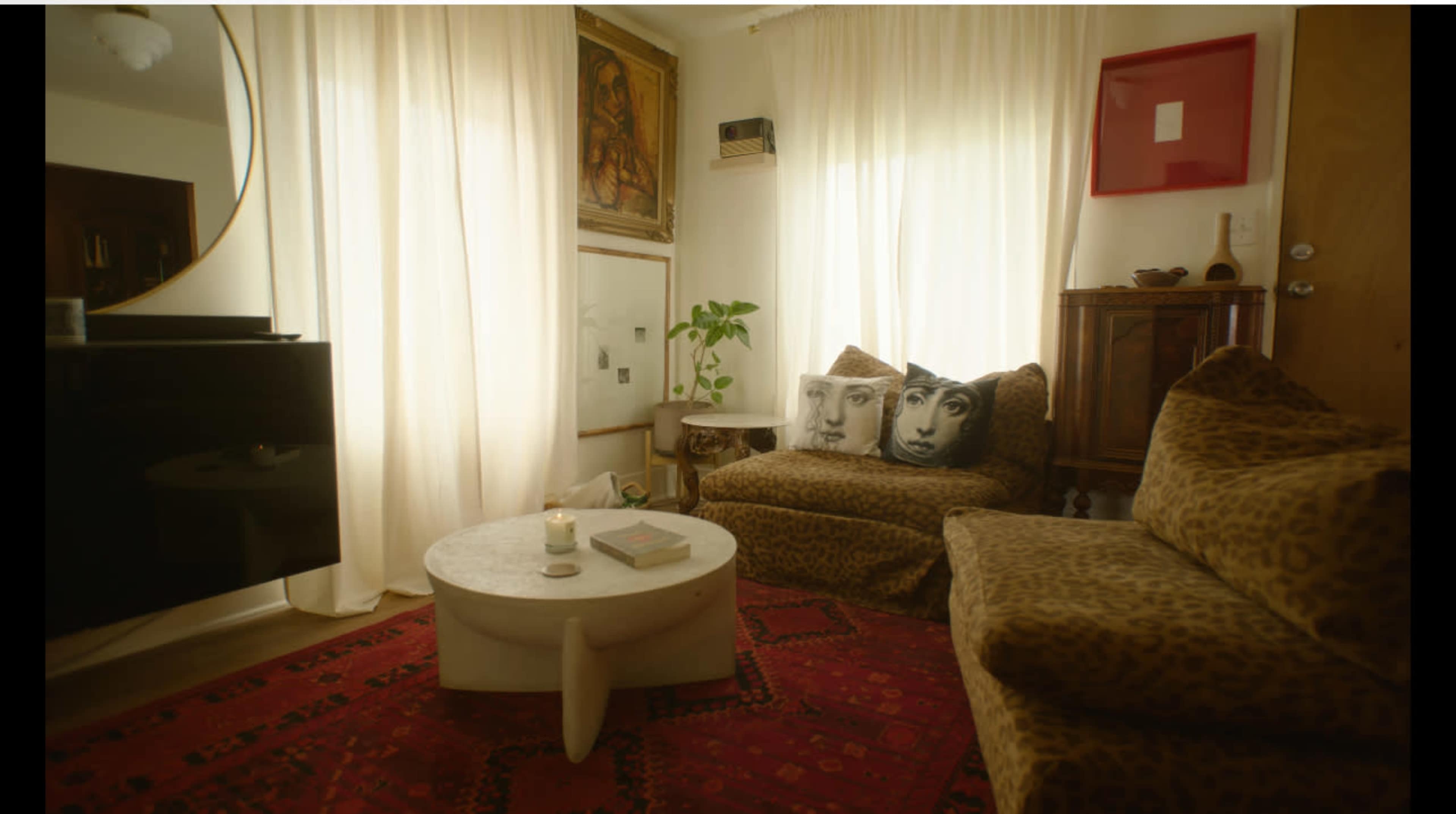 The image shows a cozy living room with leopard-print furniture, a round marble coffee table, and large windows adorned with sheer white curtains that allow natural light to filter in.