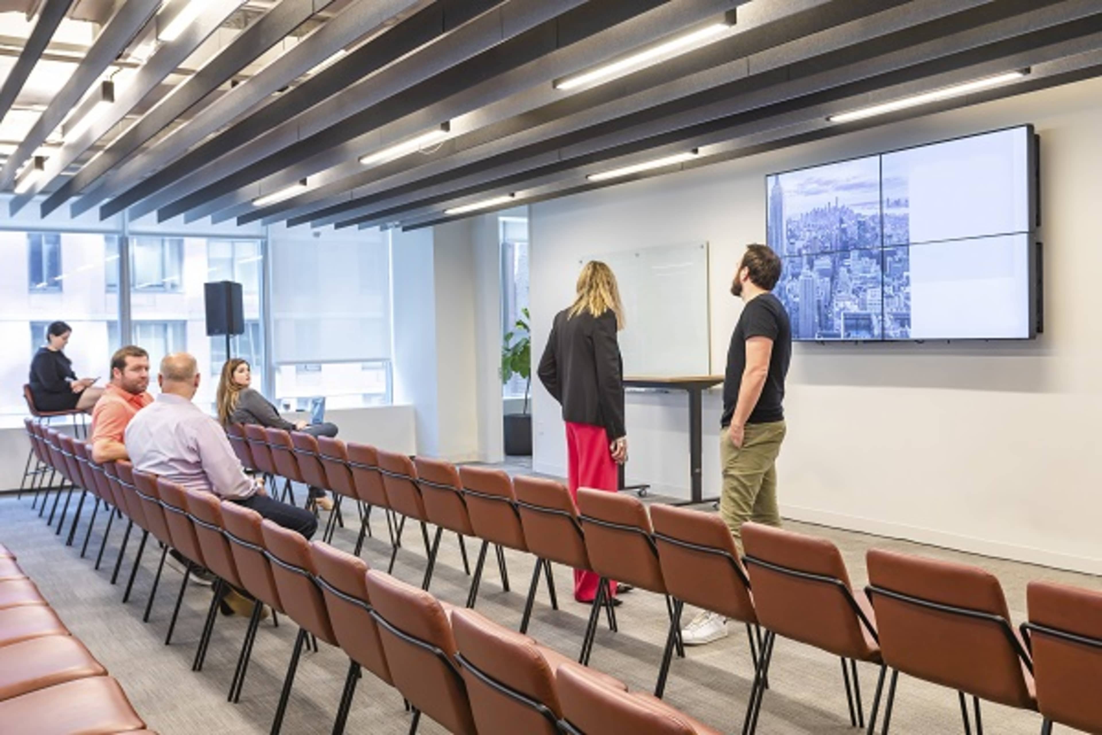 A modern conference room with rows of empty chairs facing a large screen displaying a cityscape, where two people stand near the front and others are seated in the background.